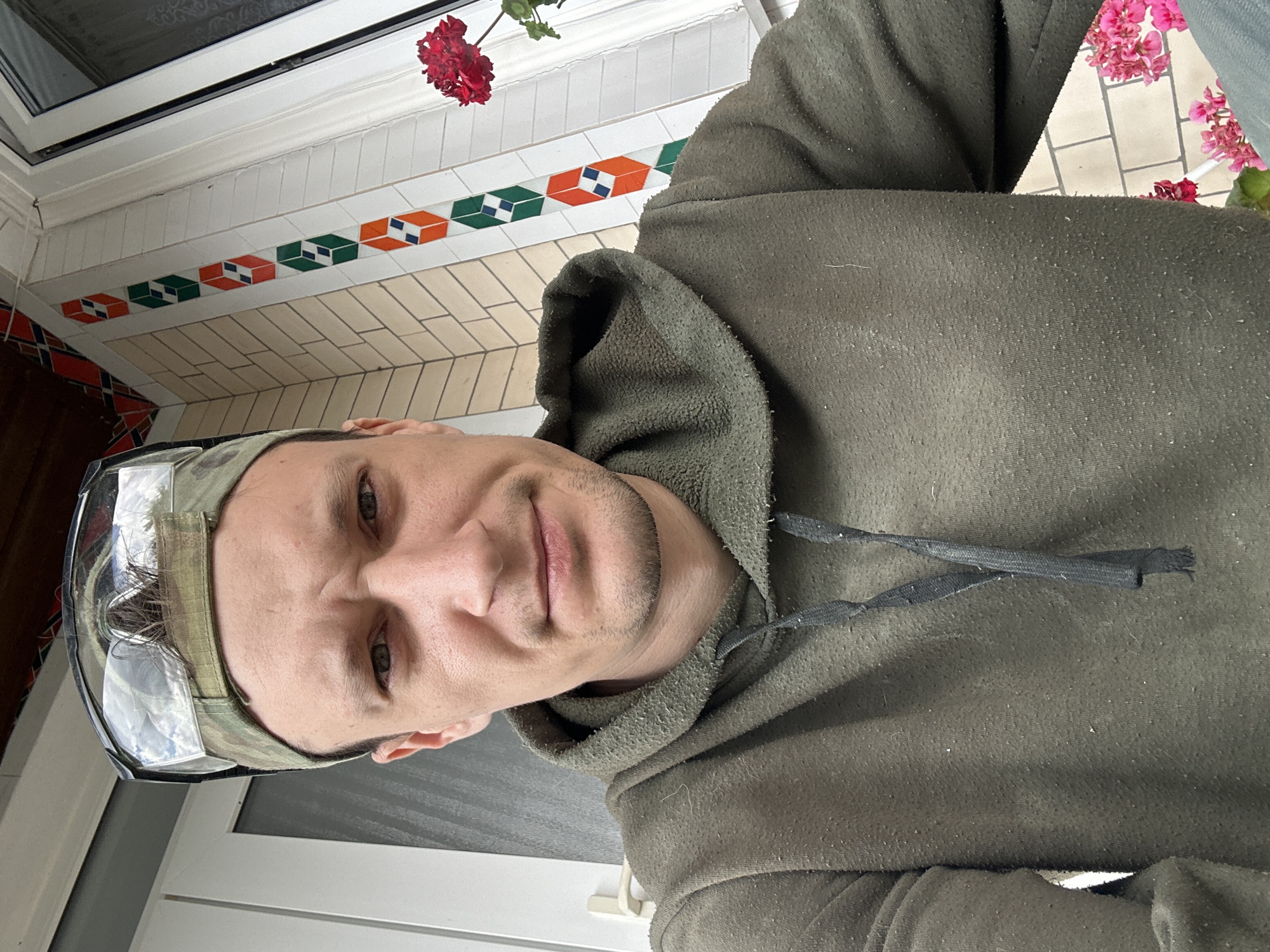 A man in a green hoodie and a camouflage baseball cap with safety glasses on top of it is taking a selfie outdoors. He has short brown hair and light brown eyes, and he is looking directly at the camera with a serious expression. The background shows a wall with decorative tiles, a window, and some pink flowers. The image appears to have been taken in Makiv, Ukraine, as indicated by the geotag.