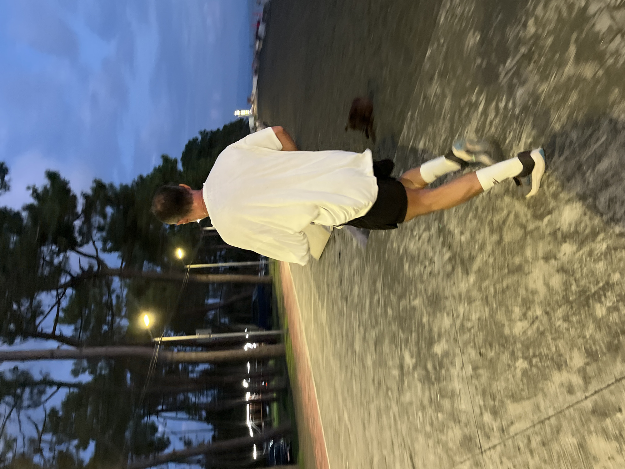 A man is captured from behind in mid-stride, running along a paved path next to a beach in Ureki, Georgia. The time of day appears to be dusk or early evening, with the sky a deep blue and streetlights illuminating the path. The man is wearing a white t-shirt, black shorts, white socks, and running shoes. The path is bordered by a red line and has a grey, textured surface. To the left of the path, a line of tall pine trees obscures a background with some lights and parked vehicles, suggesting a developed area. To the right of the path is a dark, sandy beach area, where a dark brown object, possibly a bag or clothing, lies on the ground. In the distance, the Black Sea is visible, with a lighthouse or tall structure on the horizon. The image has a motion blur, indicating the man is in motion.