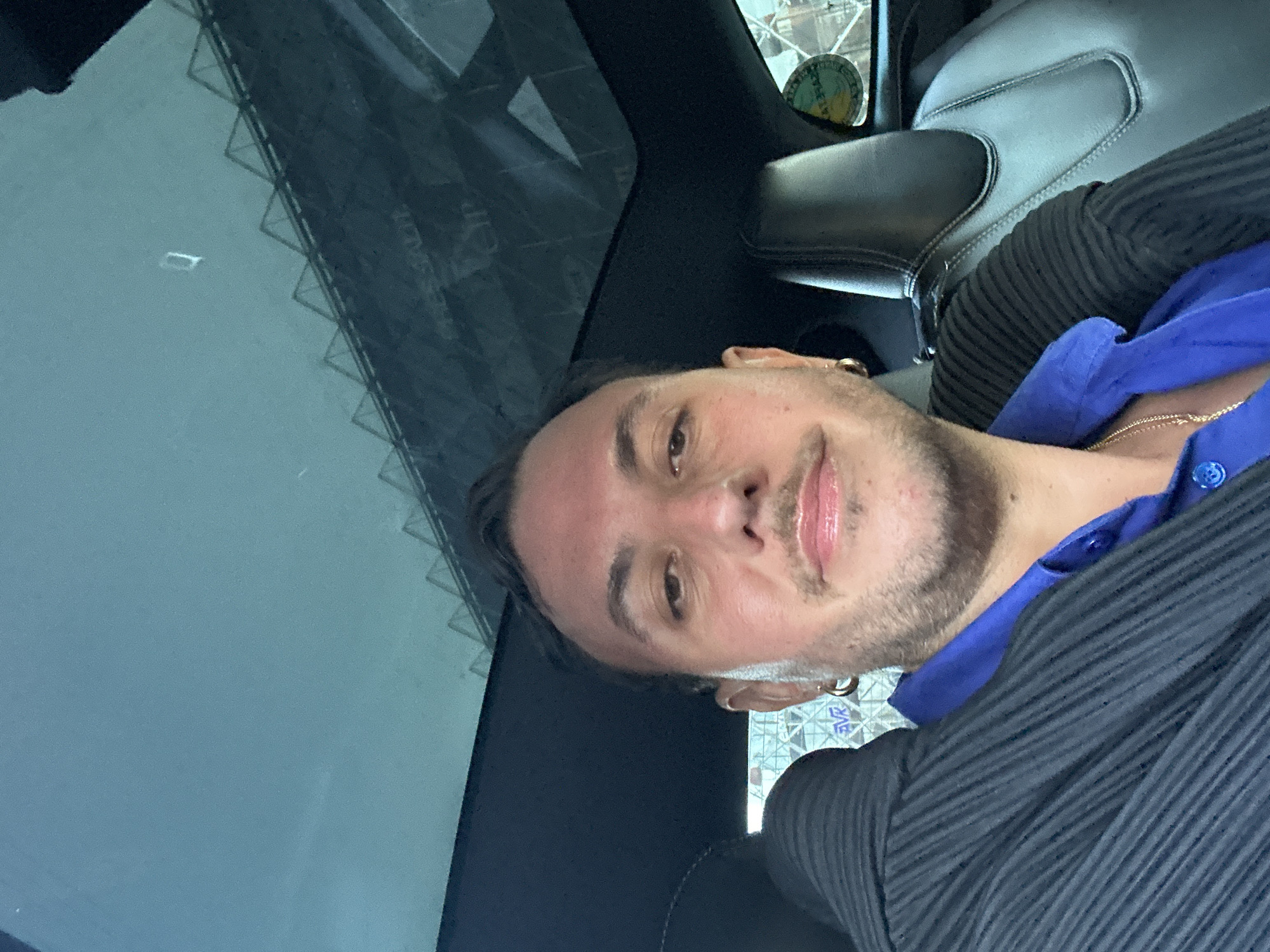 A man is in the driver's seat of a car, looking at the camera. The car has a sunroof and leather seats. The man is wearing a blue shirt and a black jacket. He has a mustache and a beard. There is a circular sticker on the window behind him. The background is blurred.