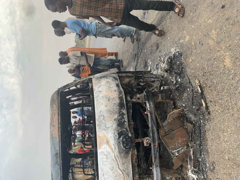 The image captures the aftermath of a vehicle fire in Alizaga, Nigeria, showing a heavily burnt-out bus or large van on a paved road. The vehicle, charred black and grey, appears skeletal, with its body panels and interior extensively damaged by flames. Twisted metal, exposed wiring, and ash are visible within its structure and on the road surrounding it.
A group of approximately ten men and boys, mostly young adults, are gathered around the vehicle, observing the damage. They are dressed in casual attire, including t-shirts, jeans, and traditional clothing. Some individuals are standing closer to the burnt wreck, looking inside or at its remains, while others stand further back. Additional, indistinct figures can be seen inside the burnt vehicle's frame.
The scene is set outdoors during daylight hours, under a hazy or overcast sky, which contributes to a somewhat muted color palette. The road surface is asphalt, and the background shows an open, relatively flat landscape. No discernible text is visible in the image. The expressions of the onlookers are not clearly visible, but their presence indicates curiosity or concern following the incident. The burnt debris on the road surface emphasizes the intensity of the fire.