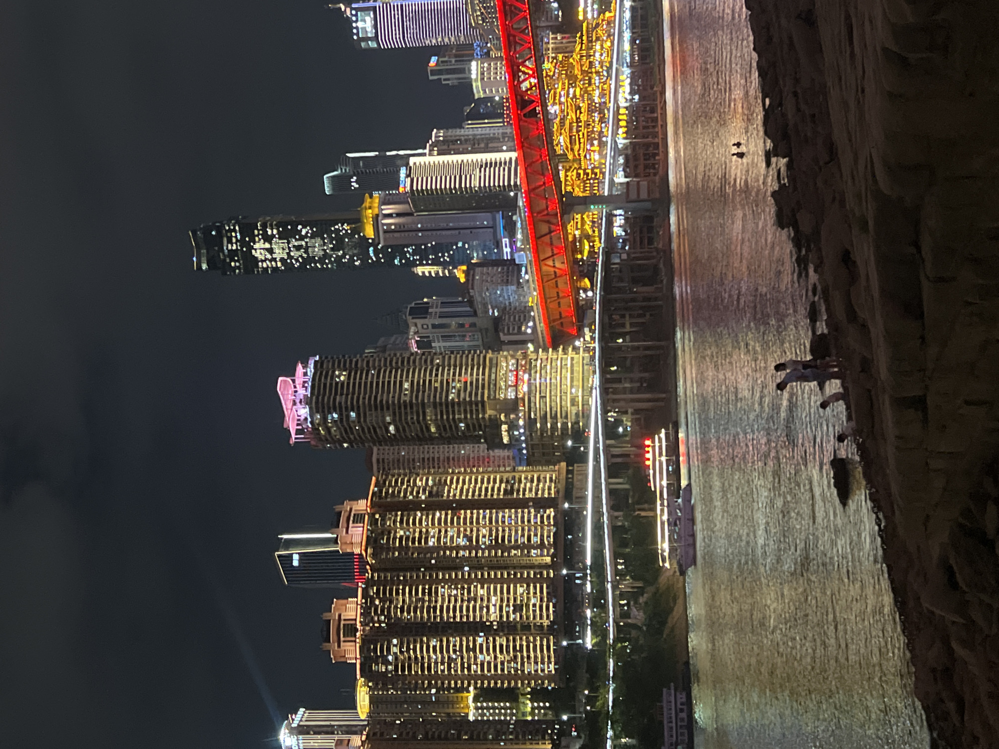 The image depicts a nighttime cityscape in Jiangbeicheng Subdistrict, Chongqing, China. A wide river occupies the foreground, with reflections of city lights shimmering on its surface. On the far bank, a cluster of illuminated skyscrapers and buildings rises against the dark sky. A prominent red pedestrian bridge spans across the river, connecting different parts of the city. Several boats are visible on the water, some docked and others moving. Along the riverbank in the foreground, a rocky shoreline is visible, with a few people standing and interacting. The overall scene conveys a vibrant urban atmosphere at night.