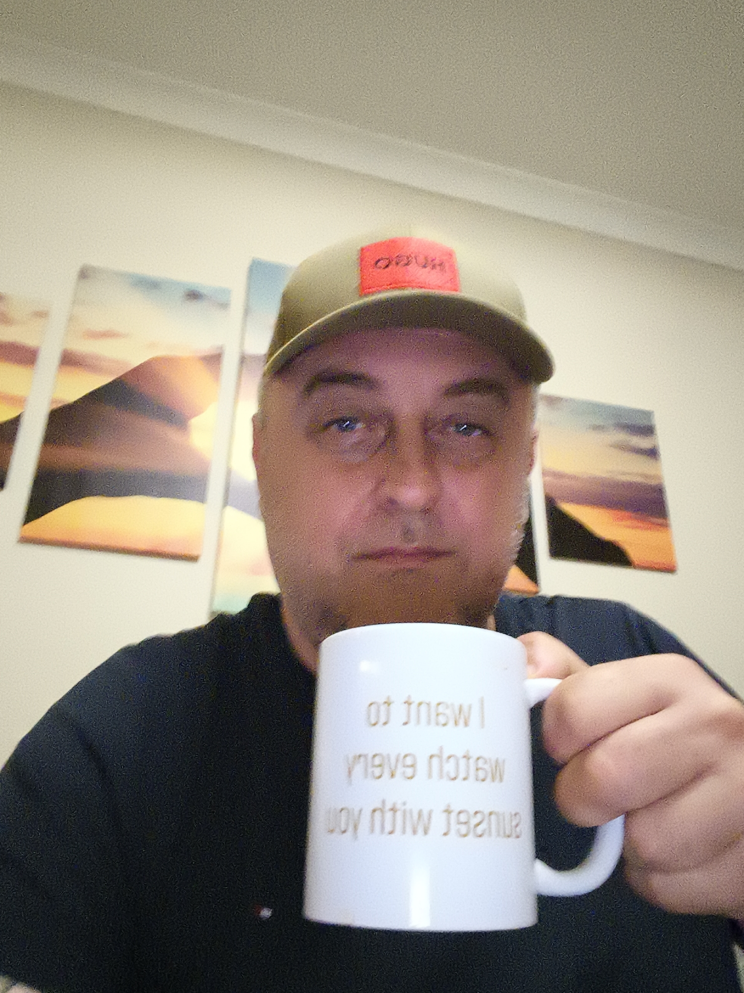 A man, appearing middle-aged, is depicted in a close-up, chest-up indoor portrait. He has a neutral expression and looks directly into the camera. He is wearing an olive green baseball cap with a prominent red square patch on the front, which displays the brand name "HUGO" (reversed in the image). He also wears a dark-colored t-shirt, possibly black or dark navy.
The man holds a white ceramic mug in his right hand, positioned in front of his chest. The mug features text printed on its side, which appears reversed due to the camera perspective. When un-reversed, the text reads: "I want to / watch every / sunset with you."
Behind the man, the background consists of a plain, light-colored wall. Hung on the wall are three framed pictures or canvases, arranged horizontally, which collectively form a panoramic image of a sunset or sunrise landscape. The artwork features warm tones of orange, yellow, and purple across a mountainous or hilly horizon. The lighting in the room is soft and even, suggesting an indoor setting under artificial illumination. The image quality is slightly grainy. The overall scene gives a sense of a casual moment indoors, located in Huntingdonshire, United Kingdom.