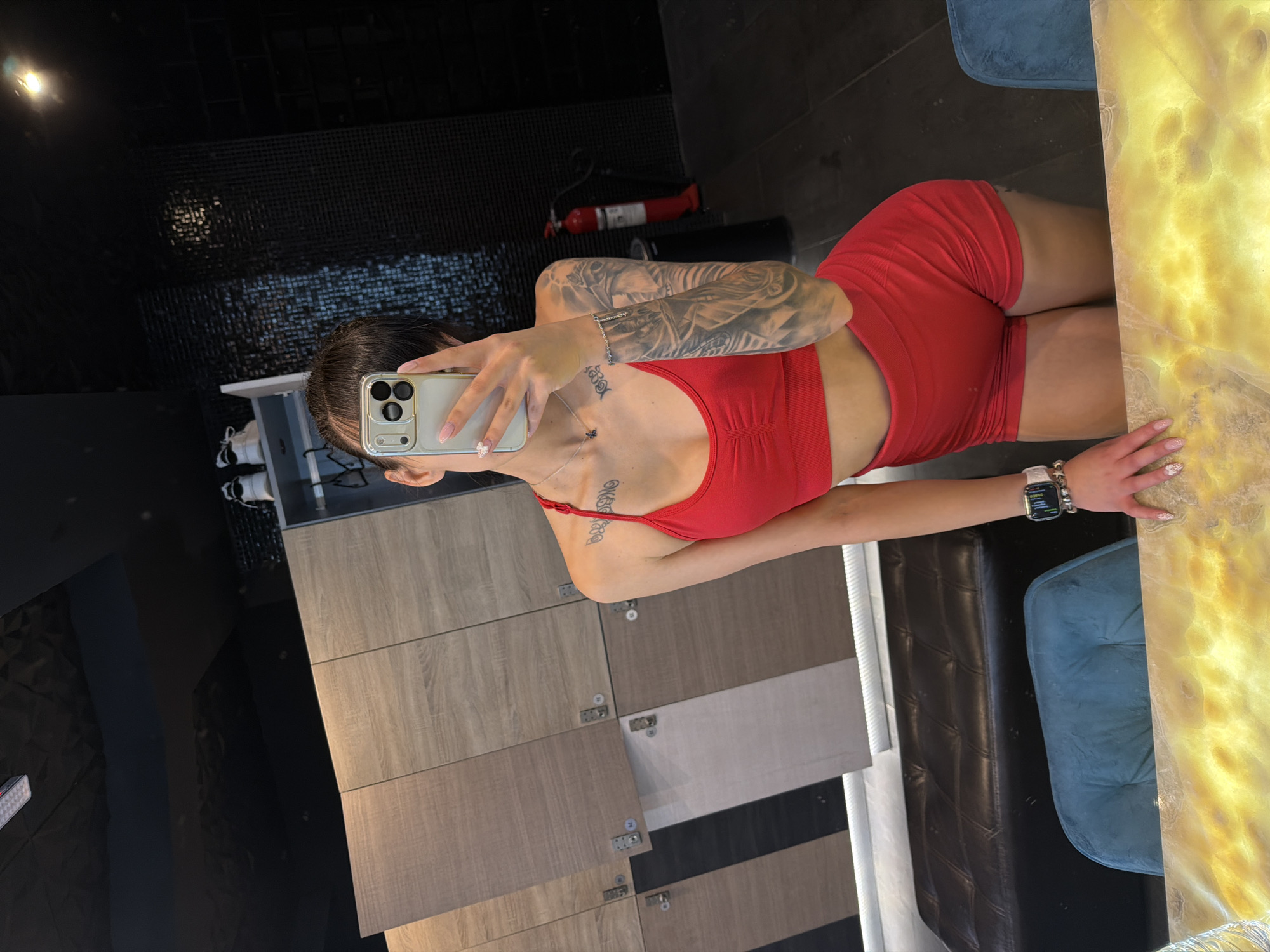 A young woman with dark hair pulled back stands in a locker room, taking a selfie in a mirror. She is wearing a red sports bra and red shorts. She has extensive tattoos on her left arm and shoulder, and a tattoo on her left collarbone. She is also wearing a watch on her left wrist and a silver necklace. The locker room features wooden lockers, a black upholstered bench, and blue chairs. In the background, a red fire extinguisher is visible. The floor is dark wood. The scene is likely from a gym or spa.