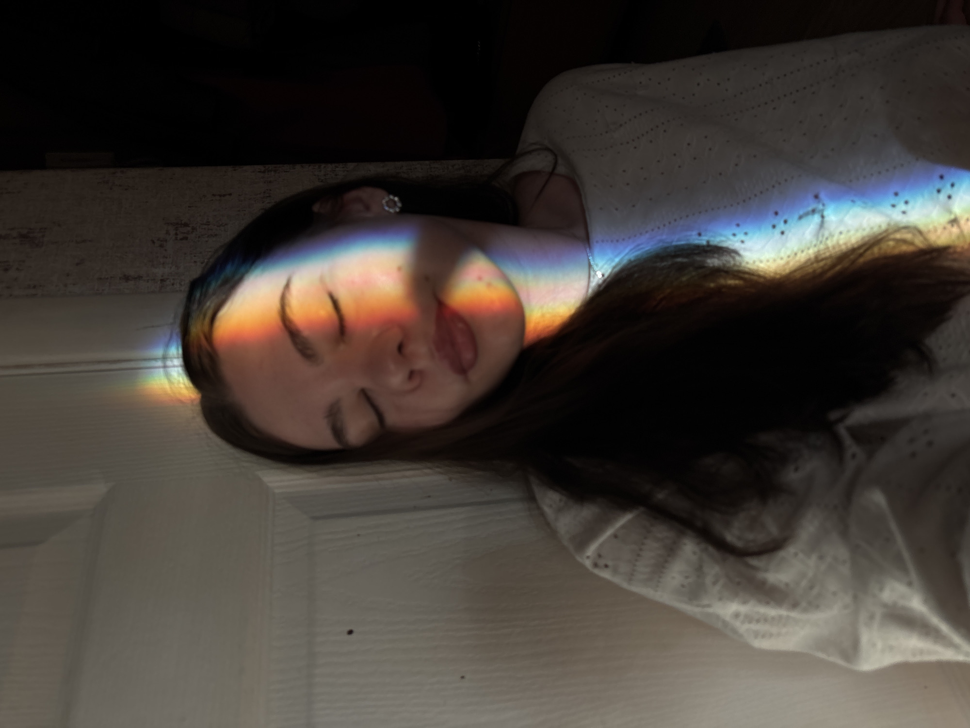 The image features a young person, likely female, with dark hair, resting or sleeping on her side. Her eyes are closed, and a serene expression is visible on her face. A vibrant rainbow light beam dramatically stretches across her face, neck, and upper chest, illuminating her features and a portion of her white garment, which appears to have an eyelet or textured pattern. A small, sparkling earring is visible in her left ear.
The setting appears to be indoors, with a white paneled door visible in the lower half of the frame and a dark, textured wall or surface in the upper left background. The overall lighting is dim, enhancing the visual impact of the colorful rainbow. The presence of the rainbow suggests it is daytime, with direct sunlight refracting through a medium (like a prism or water droplets) to create the effect. The person's relaxed posture conveys a sense of peace and tranquility. There is no visible text in the image. While the provided location context is Perm, Russia, there are no specific visual cues within the photograph to confirm this location.