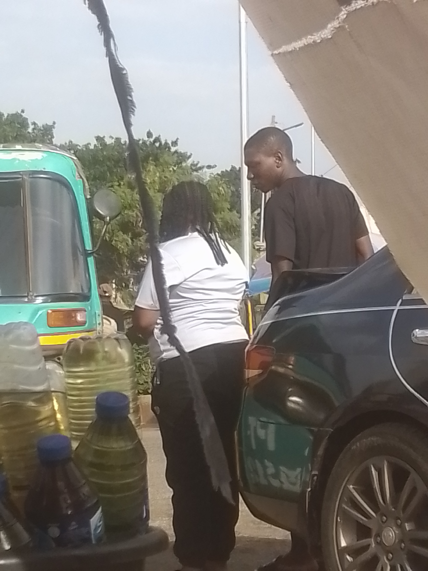 An outdoor, ground-level shot shows two individuals, a male and a female, standing near a teal-colored three-wheeled vehicle and a dark-colored sedan. The male, wearing a black t-shirt, stands on the right, facing away from the camera and looking down. The female, wearing a white t-shirt and black pants, is in the center, facing away from the camera and towards the vehicle. She has dark, braided hair. In the foreground, there are several large plastic containers, some appearing to contain liquid. The background consists of green foliage and a light-colored sky. The scene is outdoors, and the lighting suggests it is daytime. The image is framed by what appears to be a light-colored awning or structure on the right.