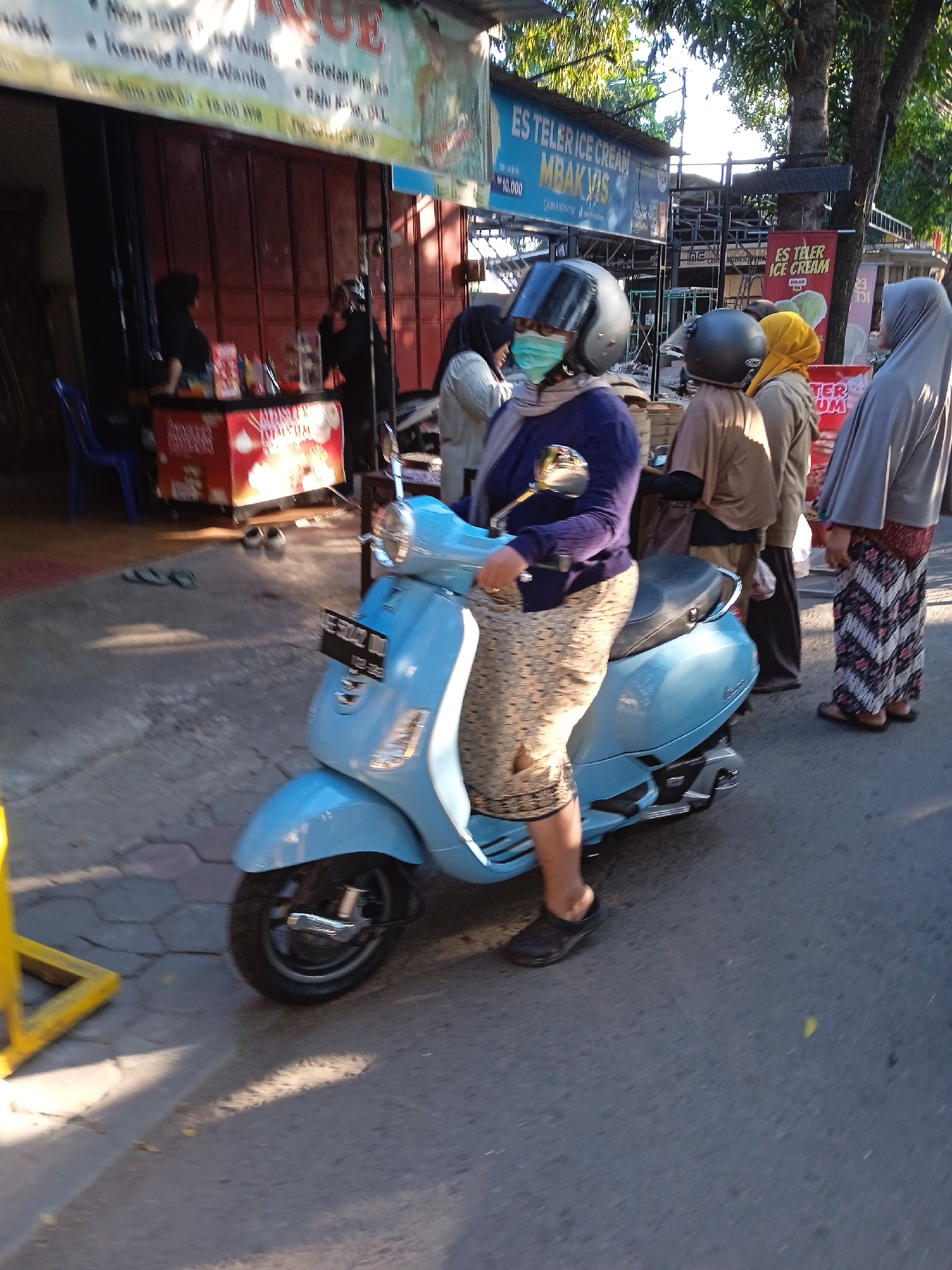 The image shows a woman wearing a helmet and a face mask on a light blue scooter, likely in Madiun, Indonesia. Several other women stand nearby. Some wear head coverings, and one wears a patterned skirt. Behind them are storefronts and food stands, including one selling "Es Teler Ice Cream." Other people are in the background. The scooter is parked on a paved surface. The light suggests it is daytime.