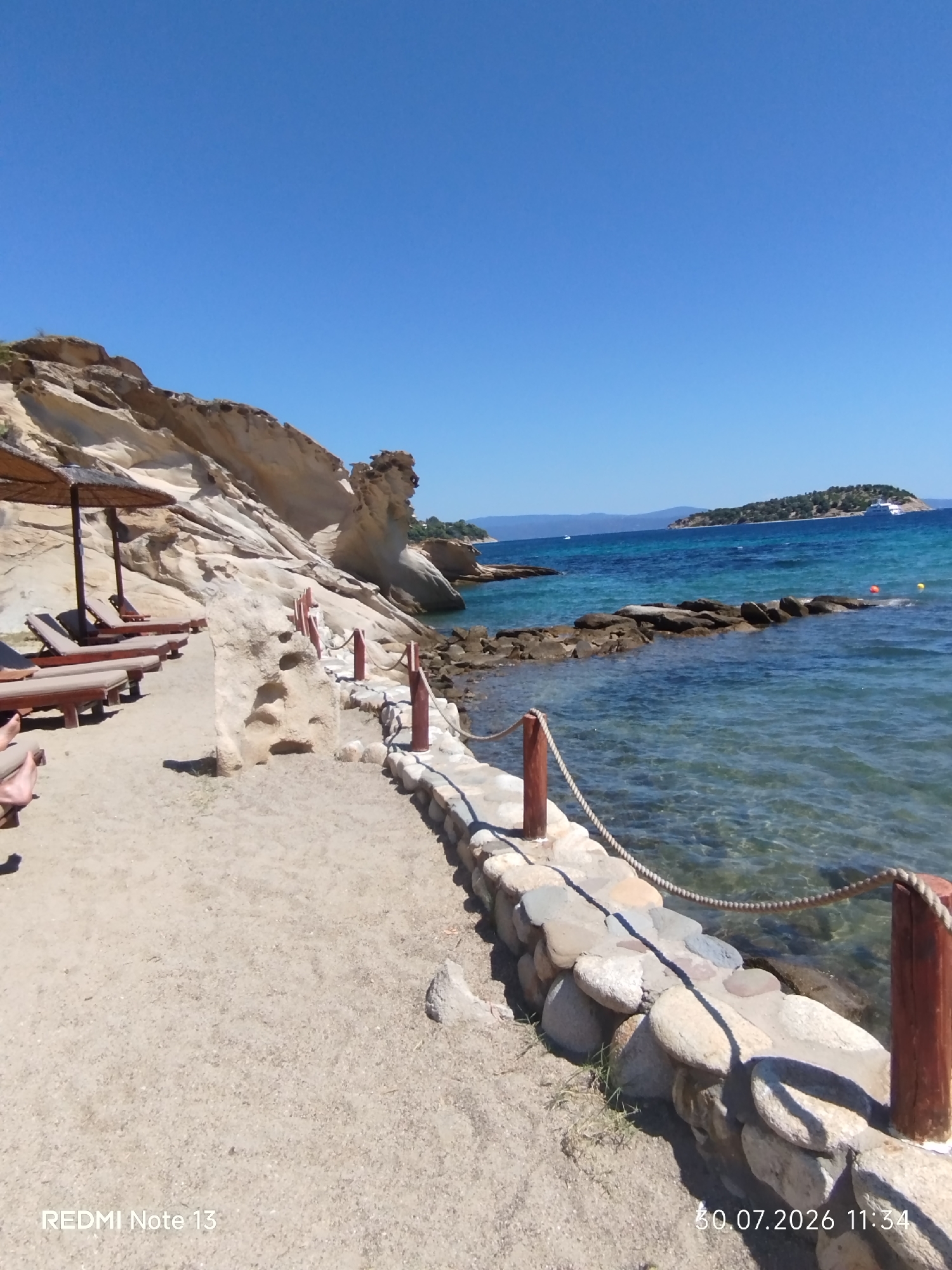 The image depicts a serene beach scene under a clear blue sky, likely in Sithonia Municipal Unit, Greece. The foreground features a sandy area with several empty lounge chairs and umbrellas, suggesting a quiet day at the beach. A stone wall, constructed with various-sized rocks, separates the sandy area from the crystal-clear turquoise water. A rope fence runs along the top of the stone wall, adding a rustic touch. In the background, a small, tree-covered island is visible in the distance, with a cruise ship docked near it. The water is calm and shallow near the shore, revealing the rocky seabed. The overall impression is one of a tranquil and idyllic coastal setting.