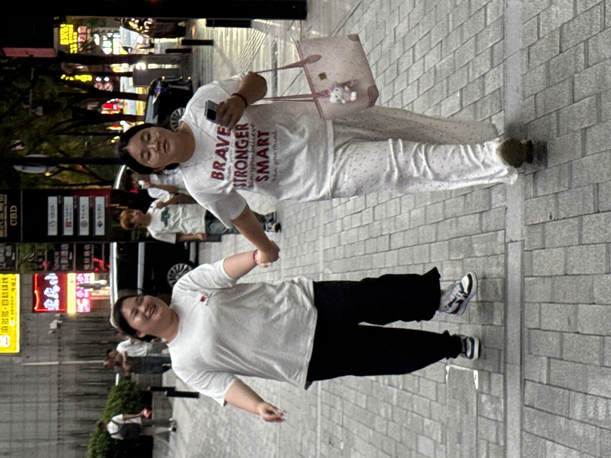 Two young women walk hand-in-hand down a paved city sidewalk in Yuzhong District, China. The woman on the left, wearing a white t-shirt and black pants, smiles broadly. The woman on the right, in white patterned pajamas and a white t-shirt with text, looks forward while holding a smartphone in her right hand and a light pink tote bag with a Hello Kitty charm in her left. The background shows a street with trees, buildings, and illuminated signs, including a yellow sign with Chinese characters and a red neon sign. Cars and pedestrians are visible in the distance. The lighting suggests it is daytime, possibly late afternoon or early evening, with some areas in shadow.