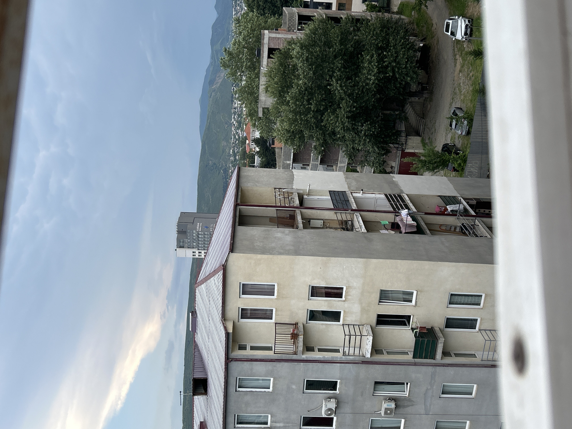 An exterior, eye-level, medium shot captures a view of residential buildings and distant mountains under an overcast sky. In the foreground, a portion of a white railing is visible. To the left, a gray apartment building features numerous windows and air conditioning units. To its right, a cream-colored building displays balconies with railings, some with laundry hanging. Beyond these buildings, a large, dense green tree dominates the mid-ground, situated next to a partially constructed building. A parking area with three cars is visible behind the tree, and further in the distance, a town with houses and hills can be seen, leading to a backdrop of layered, verdant mountains. The lighting suggests it is late afternoon or early evening.