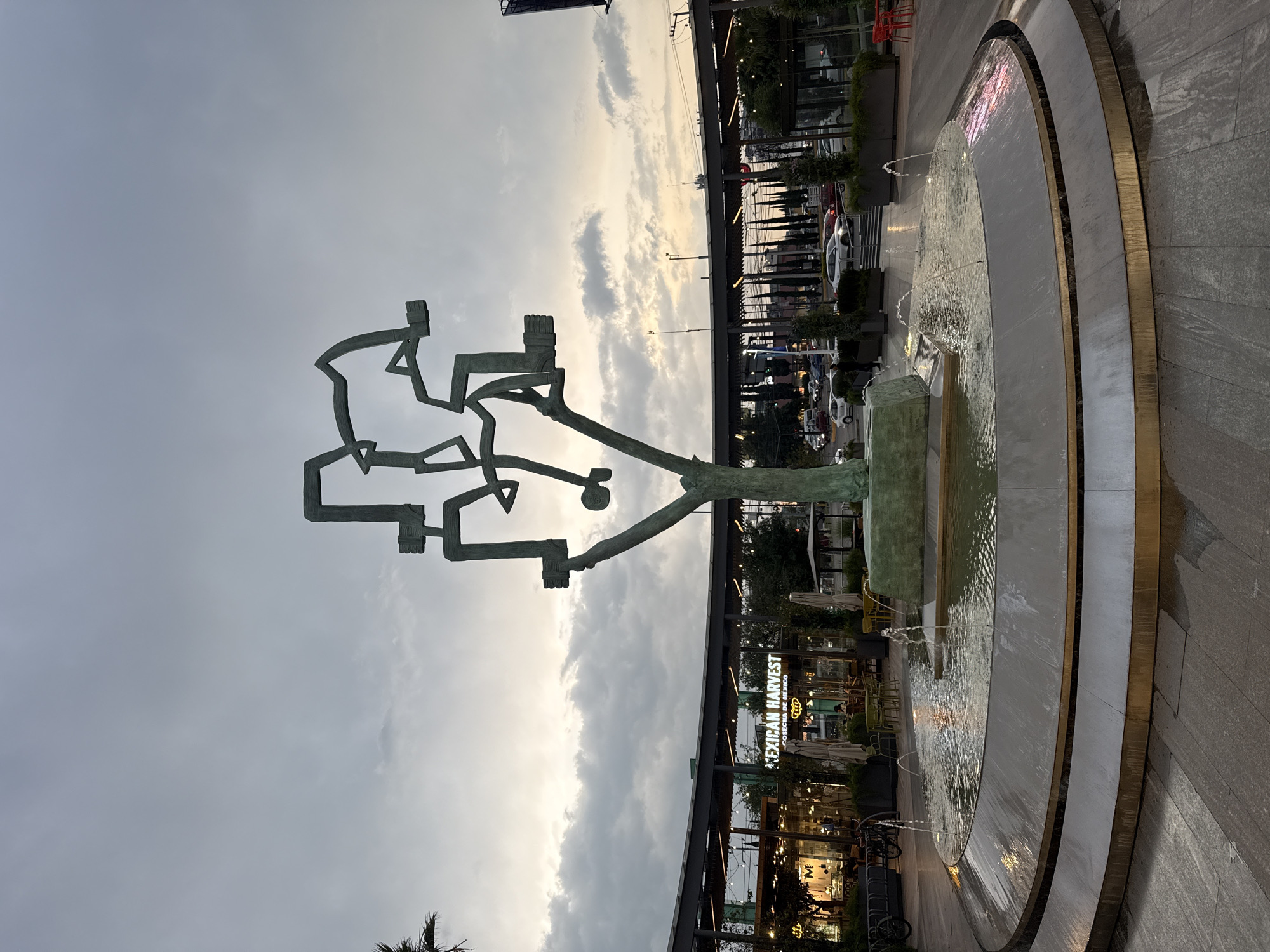 The image captures a scene in Puebla, Mexico, focusing on a public space featuring a modern sculpture and a round fountain. The sculpture, abstract in form, stands prominently against a cloudy sky. Adjacent to the sculpture is a tiered fountain with a water feature. In the background, commercial establishments are visible, including a restaurant or store with the words "MEXICAN HARVEST" prominently displayed. The surrounding area appears to be a pedestrian zone with lighting fixtures, vegetation, and some street furniture. The overall scene conveys an urban setting with elements of public art and commercial activity.