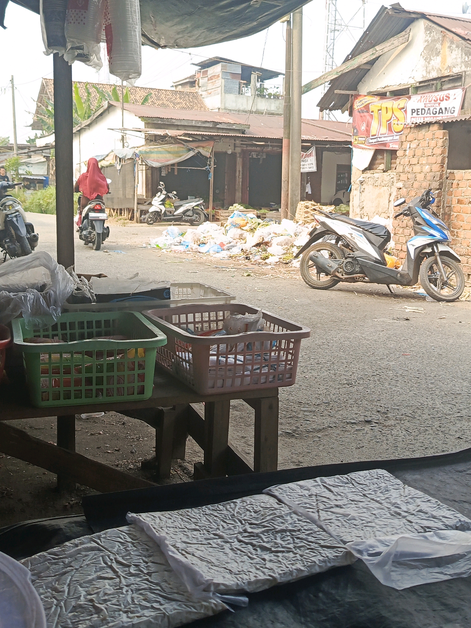 In the city of Megang Sakti I, Indonesia, a street scene unfolds in front of a market. A person wearing a red hijab is riding a motorcycle away from the viewer, partially obscured by a hanging tarp. To the right, two motorcycles are parked, one white and blue, the other white. In the background, several market stalls with corrugated metal roofs are visible, with a large pile of trash bags in front of one. A brick building with a sign that reads "TPS UNTUK PEDAGANG" (Waste Management for Traders) stands prominently. In the foreground, two plastic baskets, one green and one brown, sit on a wooden table. The ground is paved with asphalt, and the sky is overcast.