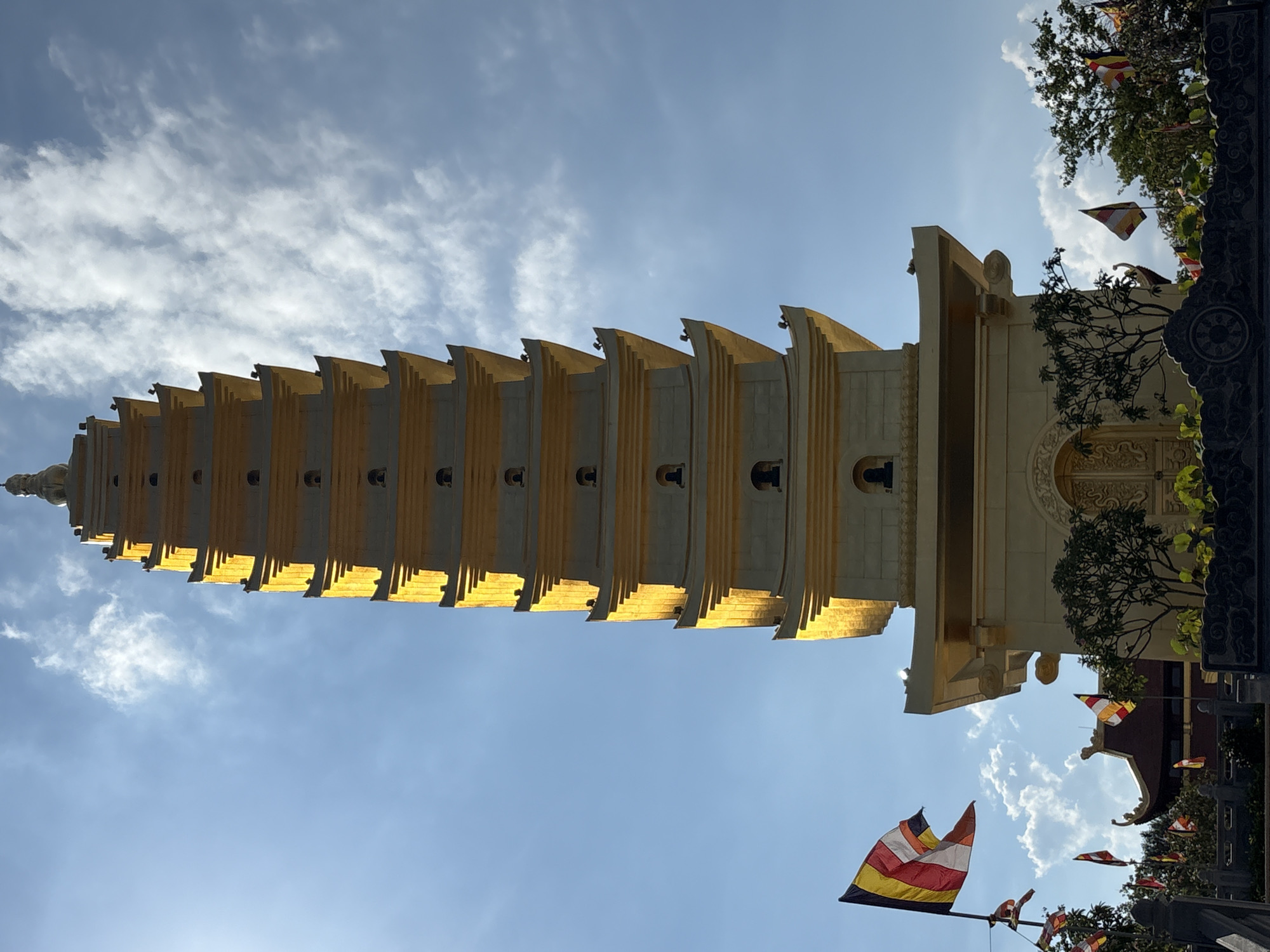 A tall, multi-tiered pagoda dominates the scene, viewed from a low angle and appearing almost vertical. The pagoda is beige and gold in color, with distinct horizontal sections and repeating architectural details. The sky is visible behind the pagoda, exhibiting a gradient from light blue to deeper blue with scattered clouds. Buddhist flags are visible near the base of the pagoda, adding to the architectural setting. The location is Diên Khánh, Vietnam, as determined by the pagoda's architectural style, the presence of Buddhist flags, and the region's typical environmental characteristics.
