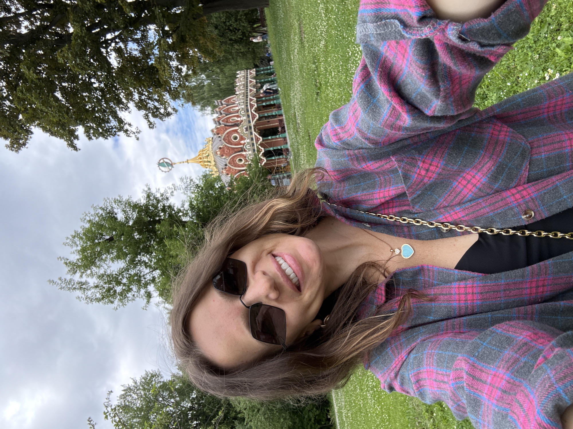 A woman is taking a selfie in front of a colorful building that resembles a carousel or pavilion in an outdoor park. She is wearing sunglasses, a plaid shirt over a black top, and a necklace with a blue heart pendant. The background shows a grassy area with scattered white flowers, trees, and a cloudy sky. The building in the background has intricate red and gold ornamentation and a unique spire. The location is the Kuskovo Estate in Moscow, Russia, known for its historical architecture and parkland.