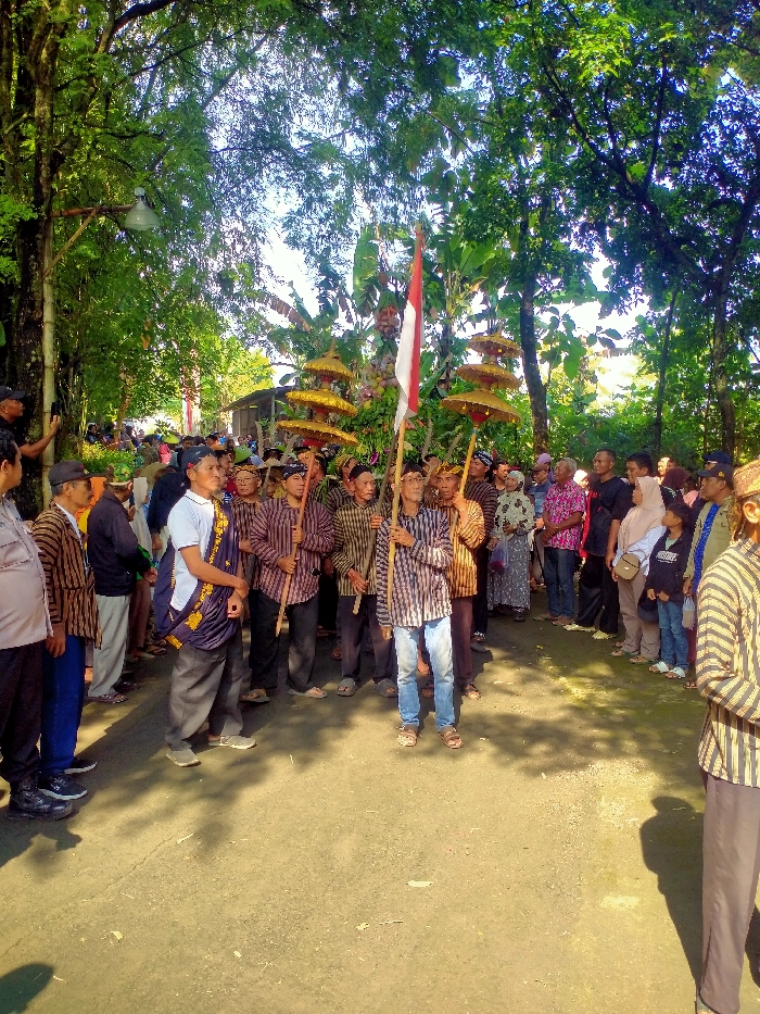 The image shows a procession of people in Masaran, Indonesia, located at the coordinates -7.471 latitude and 110.957 longitude. The group is comprised primarily of adult men, all of whom are wearing traditional Indonesian clothing, primarily brown and white striped shirts and black trousers. Some are wearing a head covering, while others are not. They are carrying wooden poles with yellow, conical-shaped umbrellas on top. The procession is being led by a man holding a red and white flag. The scene is a paved path, with trees and vegetation on both sides. There is a large group of onlookers, both men and women, some standing in the background and others seated on the ground, observing the procession. The event appears to be a traditional cultural or religious ceremony.