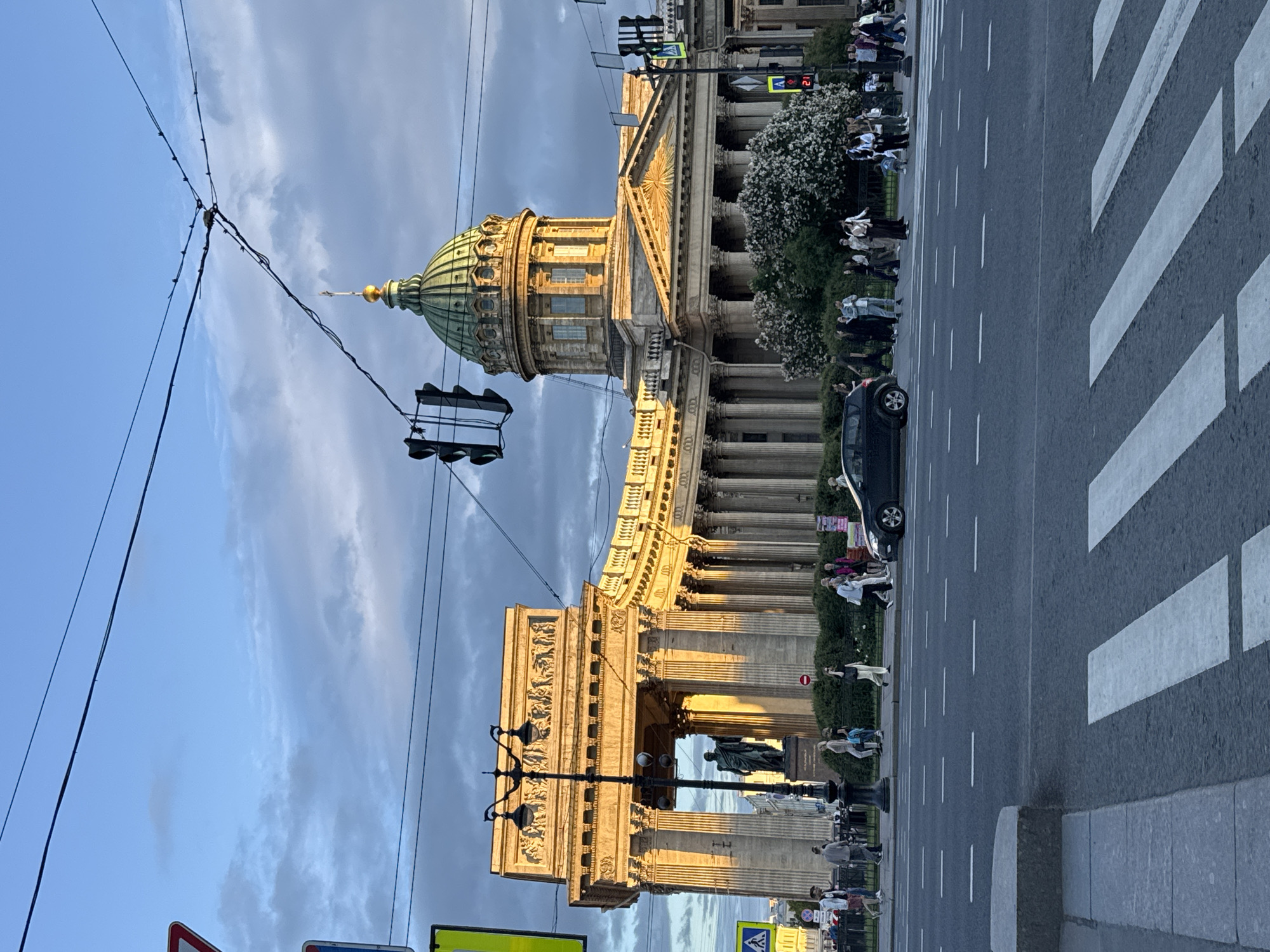 The image shows a street scene in Saint Petersburg, Russia, featuring the Kazan Cathedral. The cathedral's iconic colonnade and dome are visible, bathed in the warm glow of late afternoon or early evening sunlight. In the foreground, a pedestrian crossing is marked on the asphalt road. Several people are walking on the sidewalk in front of the cathedral, and a few cars are on the road. Traffic lights are visible, with one showing a red signal and a digital countdown. Overhead, power lines crisscross the sky, along with some clouds. The architectural style suggests the historic center of the city.