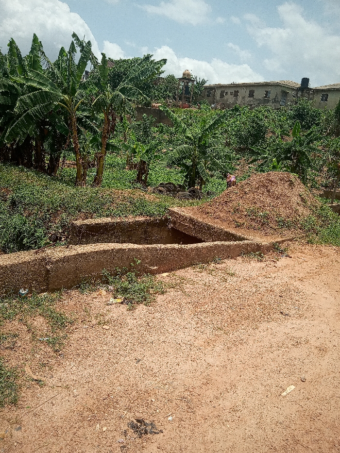 The image shows a concrete drainage ditch in a dirt lot. The ditch is partially filled with water. The ditch is located in a residential area, surrounded by banana trees and houses. The houses are made of concrete and have metal roofs. The ground is covered with dirt and some grass. There are a few pieces of trash on the ground, as well as a pile of dirt. The sky is blue and there are a few white clouds. The location is likely in the city of Ota, Nigeria. The coordinates provided are for Ota, Nigeria.