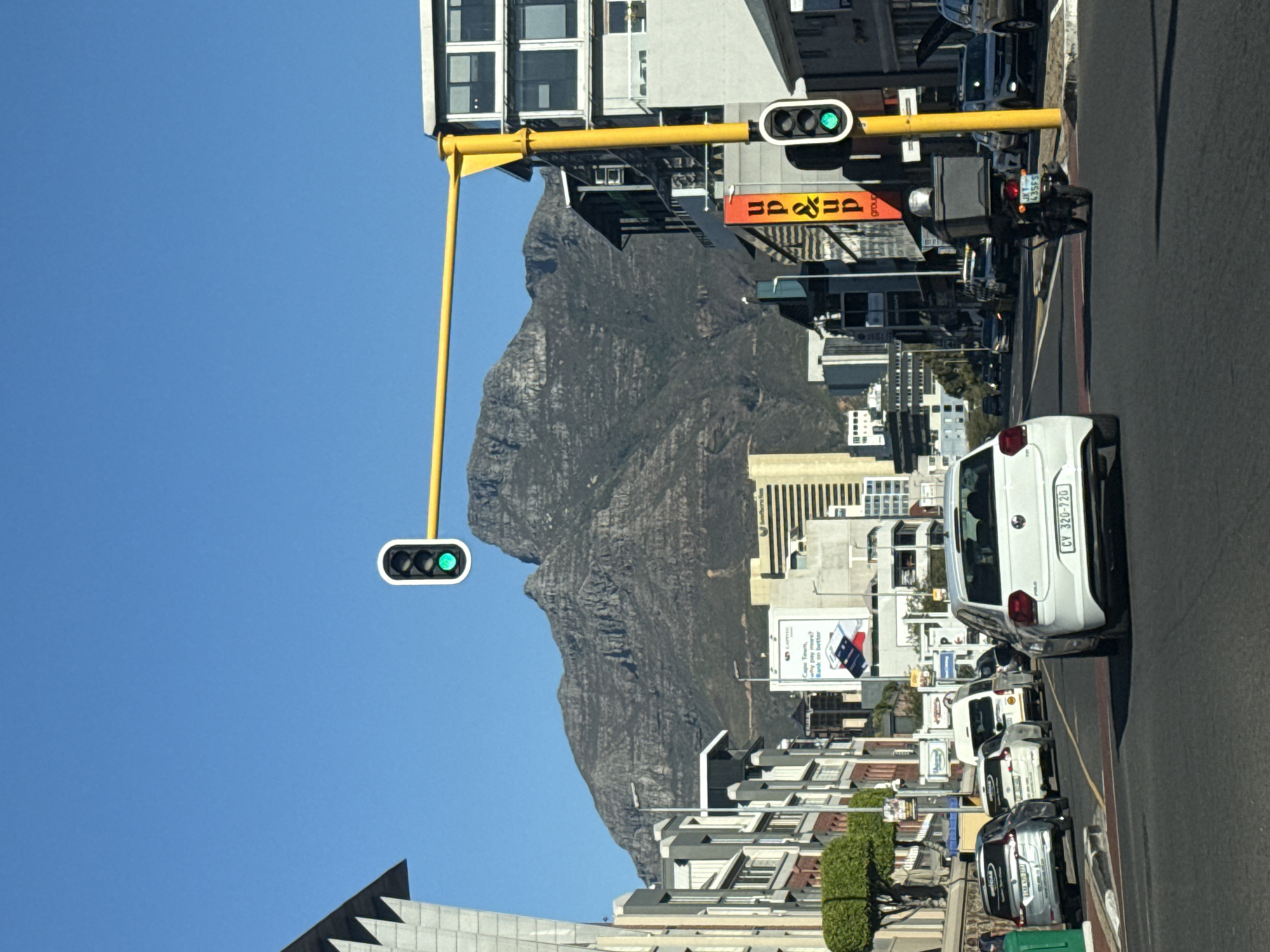 This image depicts a vibrant daytime street scene in an urban setting, with a prominent mountain in the background, likely Table Mountain in Cape Town, South Africa. The photograph is taken from a slightly elevated perspective, looking forward along a city street.
In the foreground and midground, a busy street is visible with parked cars on the right side. A white Volkswagen Polo with the license plate "CY 320-720" is closest to the viewer. Further down the street, a delivery motorcycle with a large black box on the back and a license plate ending in "435FS" is also visible. Overhead, a bright yellow pole extends across the street, supporting two traffic light units, both showing green signals, indicating that traffic can proceed.
Buildings line both sides of the street, featuring various architectural styles and multiple windows. In the midground, several signs are displayed on the buildings. One sign clearly shows "up&up group" in orange text. Another advertisement for "CAPITEC Bank" reads "Cape Town, Why pay more? Bank on better," accompanied by an image of a phone, reinforcing the location as Cape Town.
The background is dominated by the imposing, craggy slopes of a large mountain under a clear, bright blue sky, suggesting sunny weather. The overall scene captures the blend of urban life with stunning natural scenery characteristic of Cape Town.