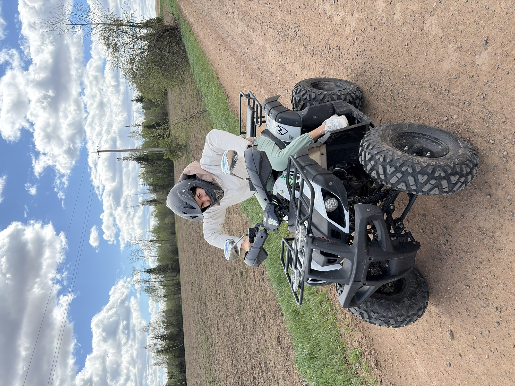 A person wearing a helmet and light-colored clothing is sitting on a black and white ATV on a dirt road next to a grassy field. The sky is blue with scattered white clouds. Trees are visible in the background. The ATV is facing towards the viewer, slightly angled to the right. The person is looking at the camera. Based on the environment, the location could be a rural area near Mozhga, Russia, given the presence of a dirt road, fields, and trees, which are common landscapes in the region.