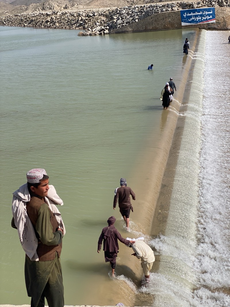 The image shows a water reservoir and dam in Kim, Afghanistan, where several individuals are walking along the dam. In the foreground, a young man with a traditional hat and clothing stands with a blanket or cloth draped over his shoulder. Downstream from him, three men are walking across the dam, with one assisting another in the water. Further along the dam, other individuals are also crossing. The reservoir is large, with rocky terrain visible on the far bank. A sign is present on the dam, with text in a language other than English.