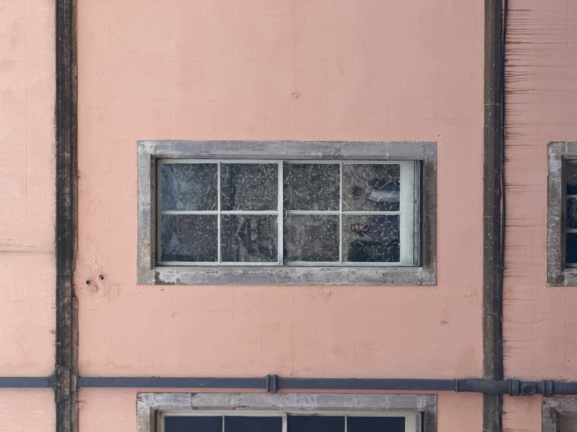 The image shows the facade of a building in an unknown city. The wall is peach-colored and composed of what appear to be square tiles. Two windows are visible, one in the center and another partially visible on the right. The central window is a rectangular shape with a gray stone frame. The window has multiple panes, and the glass is textured or patterned. The reflection in the glass is difficult to discern, but it seems to show trees or vegetation. There are vertical and horizontal dark brown architectural elements running across the facade. The vertical elements appear to be pillars, and the horizontal element looks like a pipe or beam. The wall shows signs of age and wear, with some discoloration and minor damage. The composition is symmetrical and emphasizes the architectural details of the building.