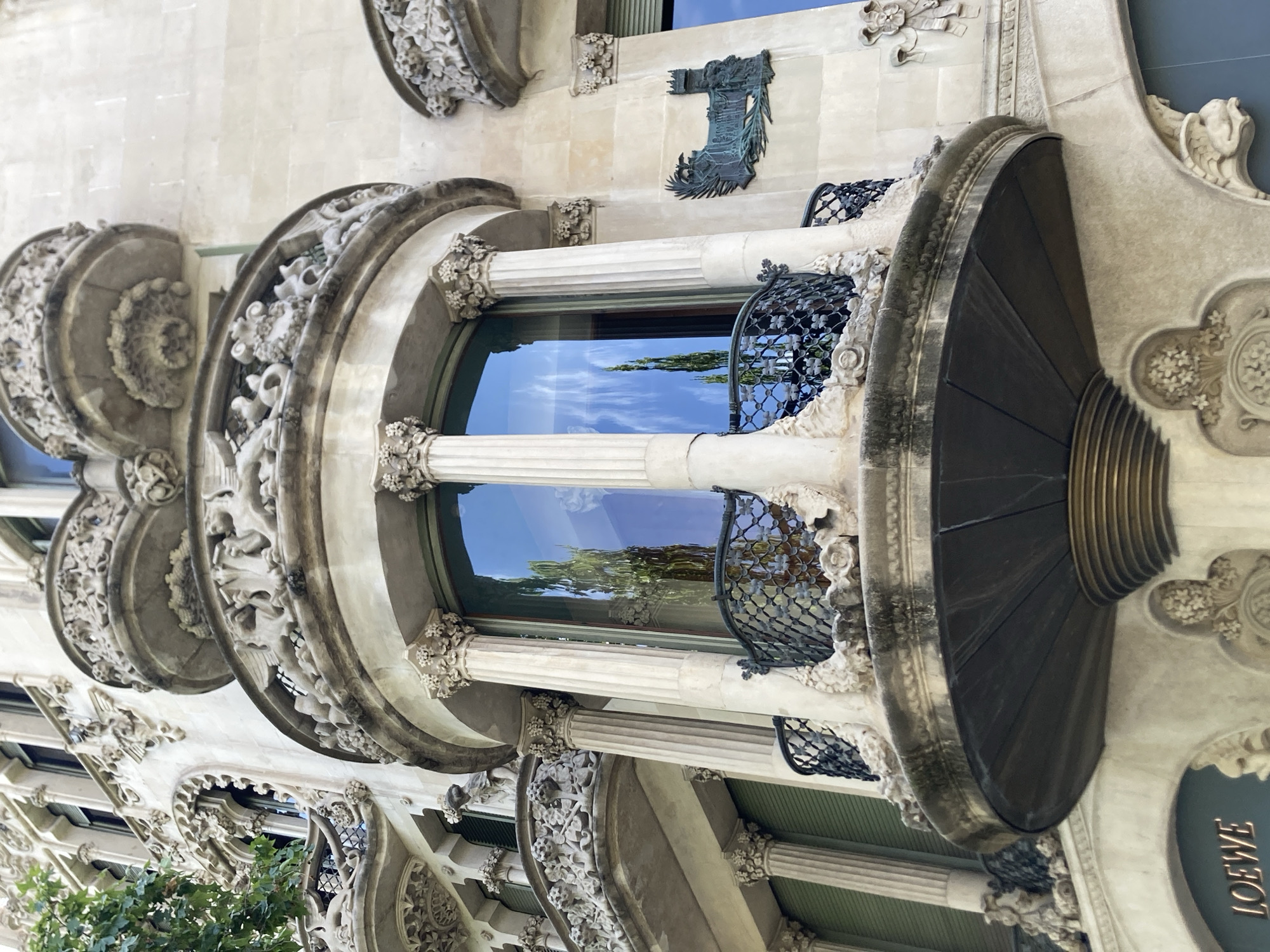 This is a photograph of the exterior of a building, likely residential, with ornate architectural details. The building's facade is made of light-colored stone and features elaborate carvings, particularly around the windows and balconies. These carvings depict floral motifs and other decorative elements.
The most prominent feature is a large, curved window that reflects a bright blue sky with scattered white clouds, suggesting it is daytime and the weather is clear. The window is flanked by fluted columns with decorative capitals. Balconies with intricate wrought-iron railings are visible, adding to the building's luxurious appearance.
Below the window, a dark, fan-shaped structure protrudes from the facade, adorned with ribbed detailing and a brass-colored rim. The overall style of the architecture is distinctive and suggests a historical period, possibly Art Nouveau or a similar movement known for its organic forms and decorative embellishments.
In the lower right corner, the word "LOEWE" is visible in gold lettering against a dark blue oval background, indicating the presence of a retail establishment or a branding element on the building. Green foliage from trees is visible in the lower left portion of the image, suggesting the building is situated in an area with greenery. The image is oriented vertically, with the top of the building towards the left of the frame. The scene evokes a sense of grandeur and artistic craftsmanship.
