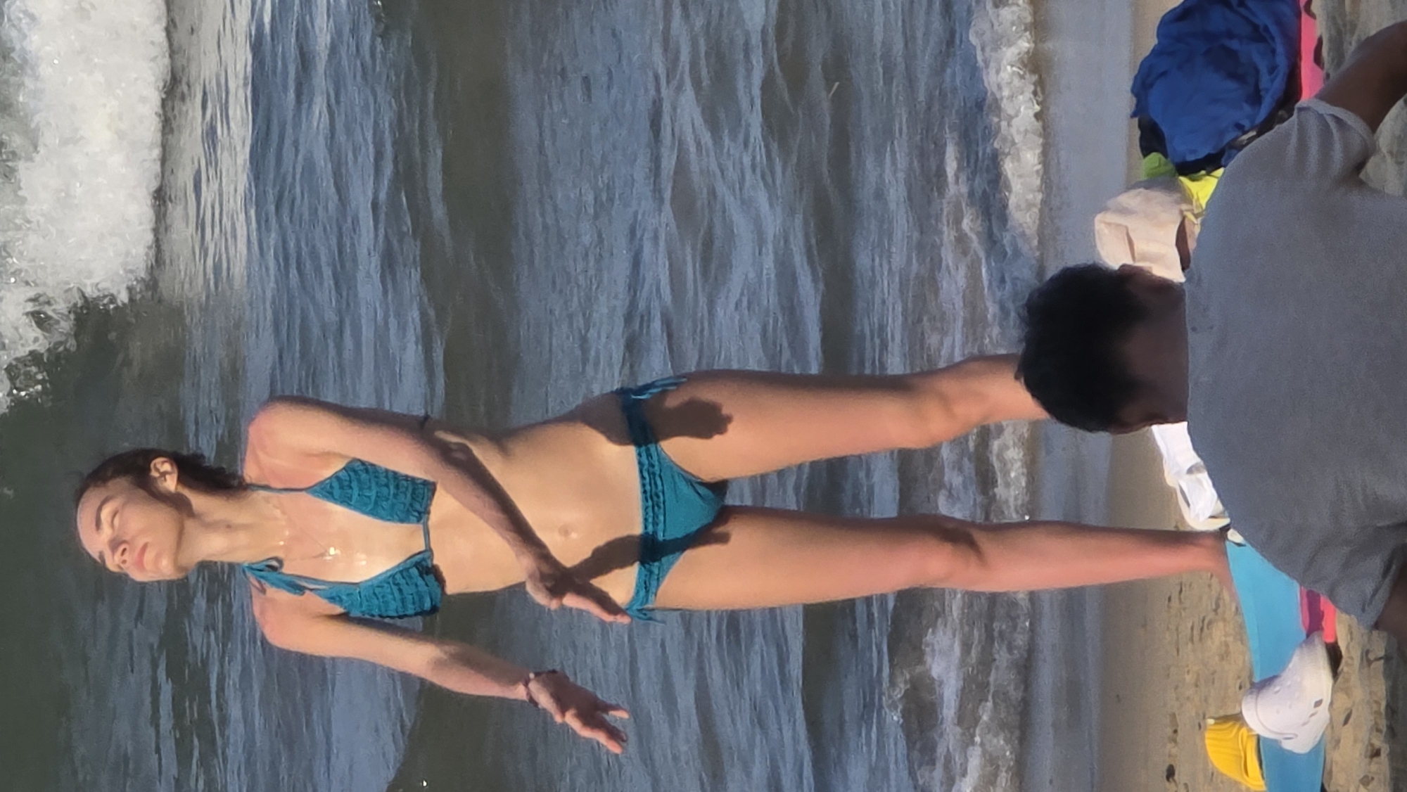 A woman in a blue bikini stands in the water at a beach. To the right, a person stands on the sand, wearing a grey shirt. The scene appears to be set in Nha Trang, Vietnam, based on the characteristics of the beach and water.