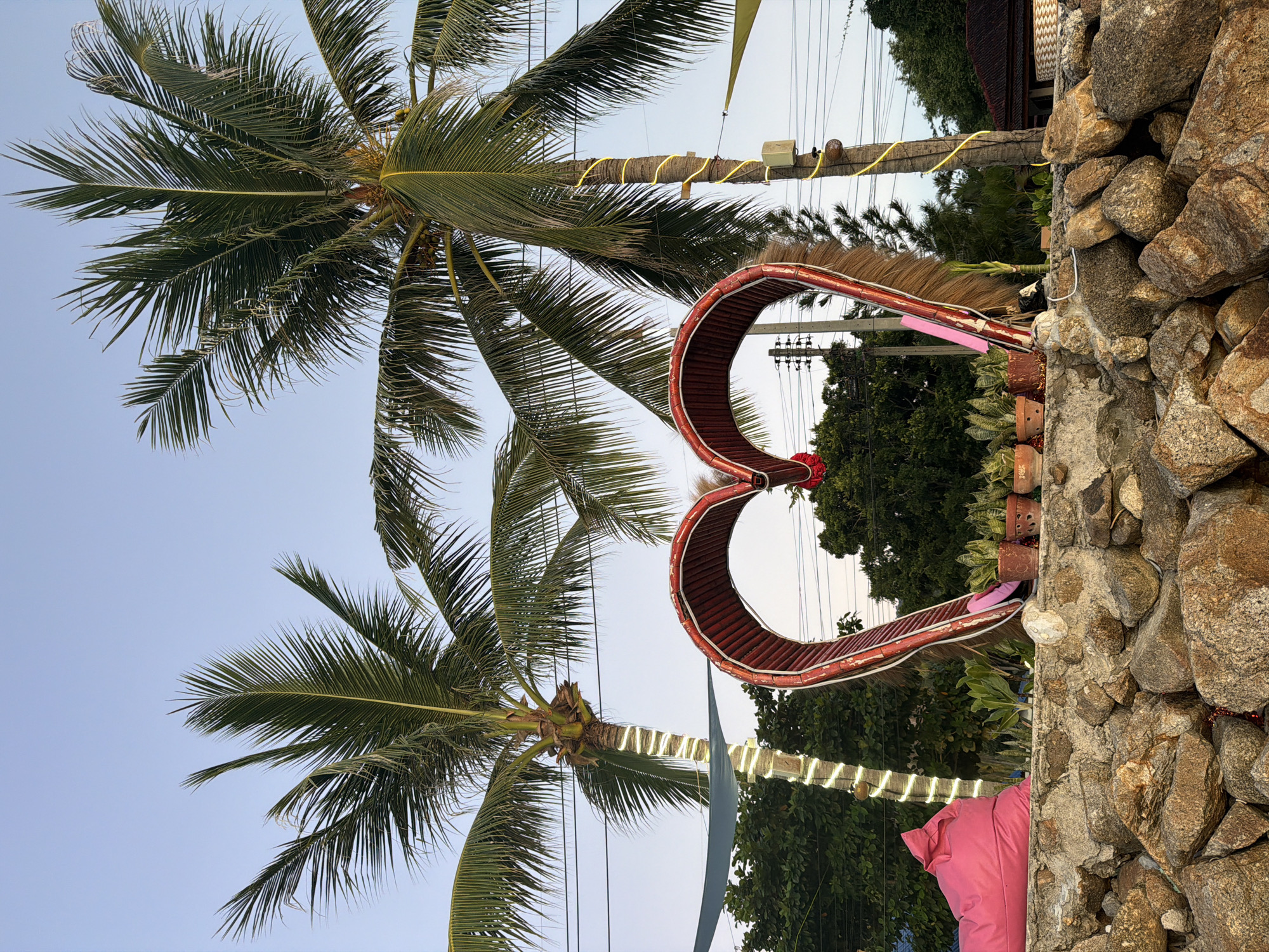 In Ko Pha Ngan, Thailand, a large, red, heart-shaped archway is positioned on a stone wall. Two palm trees flank the heart, with one adorned with white string lights and the other with yellow string lights. A bright pink beanbag chair sits to the left of the heart, and several potted snake plants are placed in front of it. The scene is set against a pale blue sky.