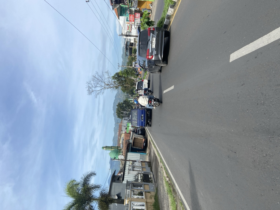 The scene is a street in Soreang, Indonesia, at latitude -7.032, longitude 107.528. The road is paved and has a white lane divider. There are a few cars and motorcycles traveling in both directions. There are buildings and trees along the sides of the street. The sky is blue and clear. There are power lines and a few electrical poles along the street. In the distance, a green-domed mosque can be seen.  A person is riding a motorcycle in the middle of the lane, with the other motorcycle and vehicles following behind at a safe distance. The person is looking forward at the road. The environment is mostly residential and suburban, with small buildings and houses, trees, and a few shops or businesses.