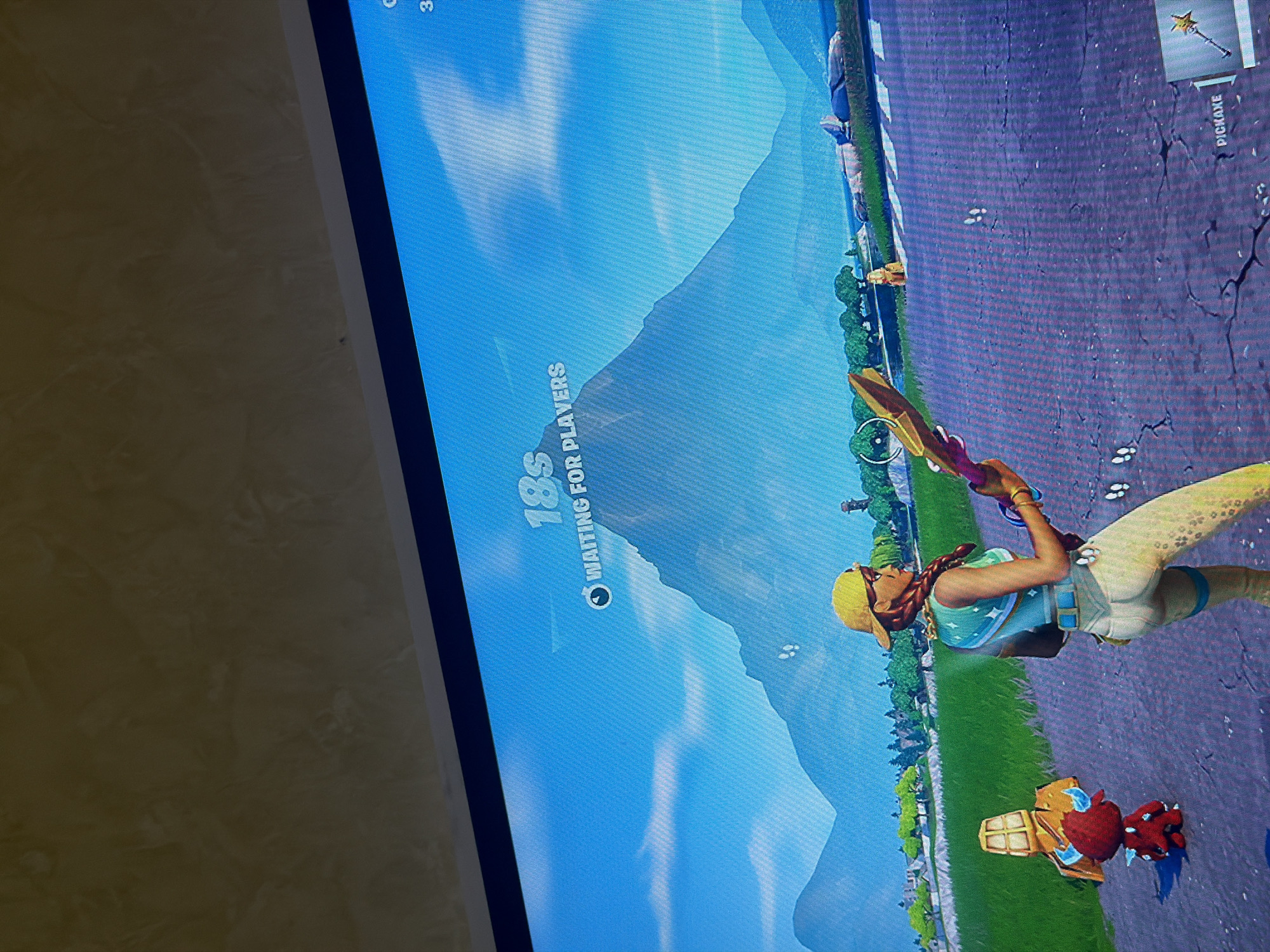 This media file, taken in Iskar, Bulgaria, presents a vertically oriented photo of a screen displaying a video game scene, likely Fortnite, rotated 90 degrees counter-clockwise.
The primary subject is a female game character, light-skinned with braided hair, wearing a yellow cap, a light blue top with a star design, and light-colored shorts or pants. She holds a golden pickaxe over her shoulder with her right hand. On the cracked asphalt road surface near her feet, a red object resembling an apple is visible next to a golden, block-like structure. The road is bordered by a strip of green grass and trees. In the background, large, stylized mountains rise under a clear, bright blue sky, indicating a daytime setting.
The scene depicts a pre-game lobby or waiting area. Text overlaid on the screen reads "18S" (a countdown timer) and "WAITING FOR PLAYERS," indicating the current game state. In the top right corner, the word "PICKAXE" is visible, likely identifying the selected in-game tool. A partial number "3" can also be seen in the upper left corner. The image shows typical screen pixelation and the bezel of the display on the left edge.