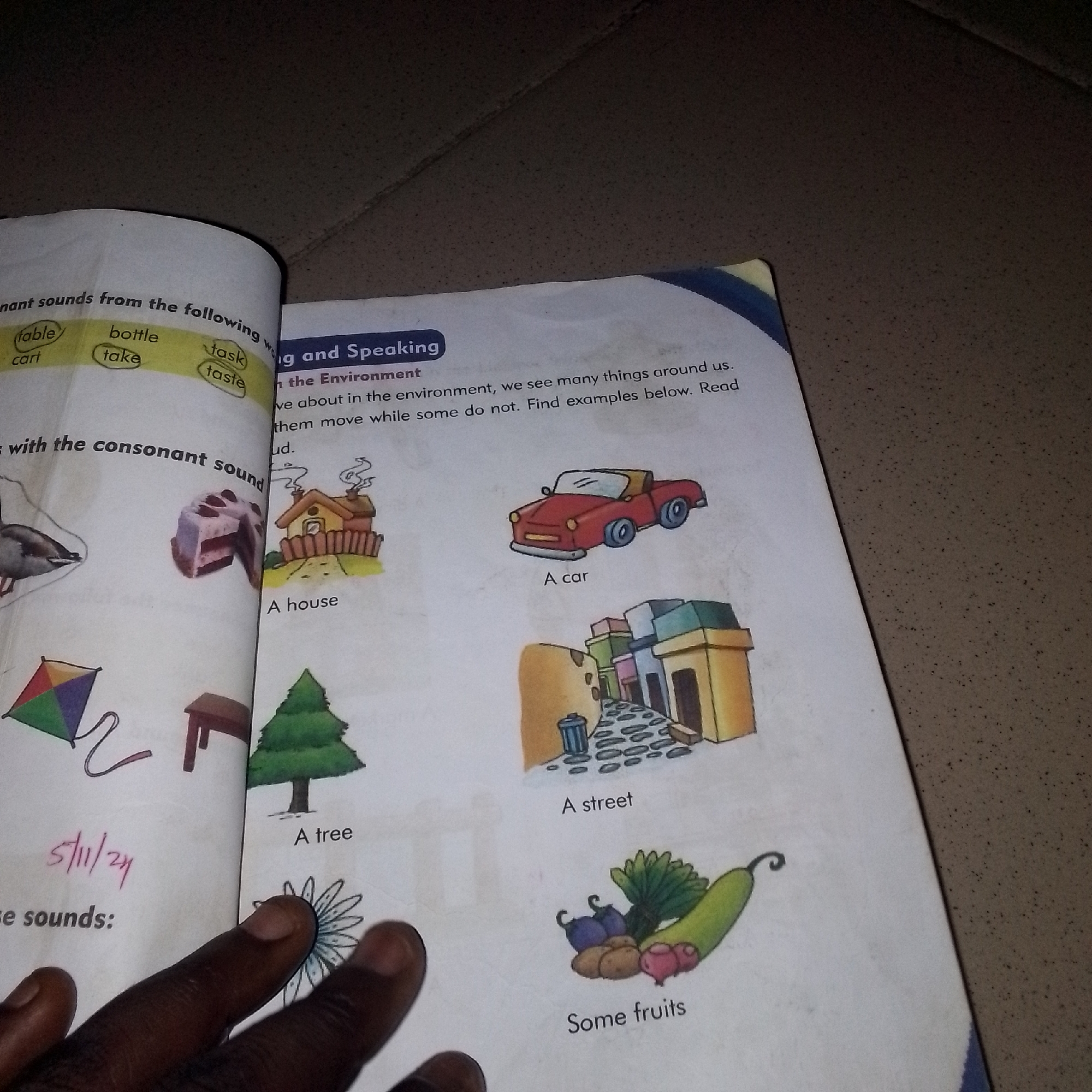 The image shows an open book on a textured surface. The left page of the book has a heading "nant sounds from the following" with words like "table", "bottle", "cart", "take", "task", and "taste" circled. Below this, there's a drawing of a goose, a slice of cake, and a kite. The right page has a heading "g and Speaking" followed by text related to the environment, including "in the Environment". Below this, there are several illustrations: a house, a car, a street scene with buildings, a tree, and a collection of vegetables and fruits. A human hand is visible in the foreground, touching the illustration of a flower. The book is open, suggesting it is being used for reading or learning. The background appears to be a dark, speckled surface, possibly a table or floor. There is no information in the image or any context provided that would suggest the location is Jalingo, Nigeria.