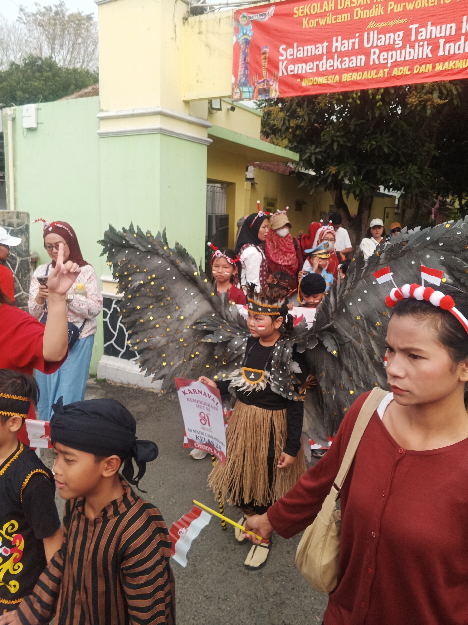 In the city of Sokanegara, Indonesia, an outdoor procession is taking place in front of a building with a light green facade and a cream-colored overhang. A large red banner is prominently displayed above the entrance, reading "Selamat Hari Ulang Tahun Kemerdekaan Republik Indonesia" (Happy Indonesian Independence Day). The banner also features text in smaller font indicating "Sekolah Dasar Negri Korwilcam Dindik Purwokerto" and "Indonesia Berdaulat Adil dan Makmur." A group of people, including children dressed in celebratory costumes, are participating in the event. One child is adorned with large, dark, feathered wings decorated with gold sequins. Another child wears a traditional striped shirt and a black head covering. A woman in the foreground wears a red cardigan and a beige shoulder bag. Several individuals are holding Indonesian flags, which are red and white. The scene suggests a community celebration for Indonesia's Independence Day.