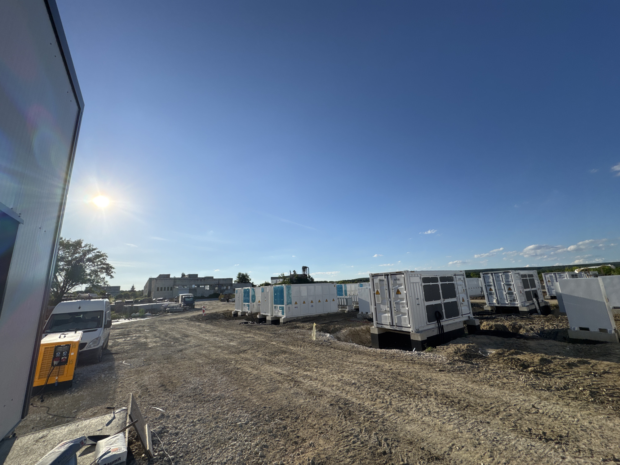 In Dolni Chiflik, Bulgaria, a construction site is visible under a clear blue sky, with the sun shining brightly. The scene features several large, white shipping containers with blue accents, arranged in rows on a dirt and gravel surface. These containers appear to be part of an energy storage system, with some displaying hazard symbols and serial numbers.
In the foreground, a white van is parked next to a yellow generator. The ground is uneven, indicating ongoing construction work. In the background, industrial buildings and trees are visible. The overall impression is one of a developing infrastructure project in a semi-urban or industrial area.