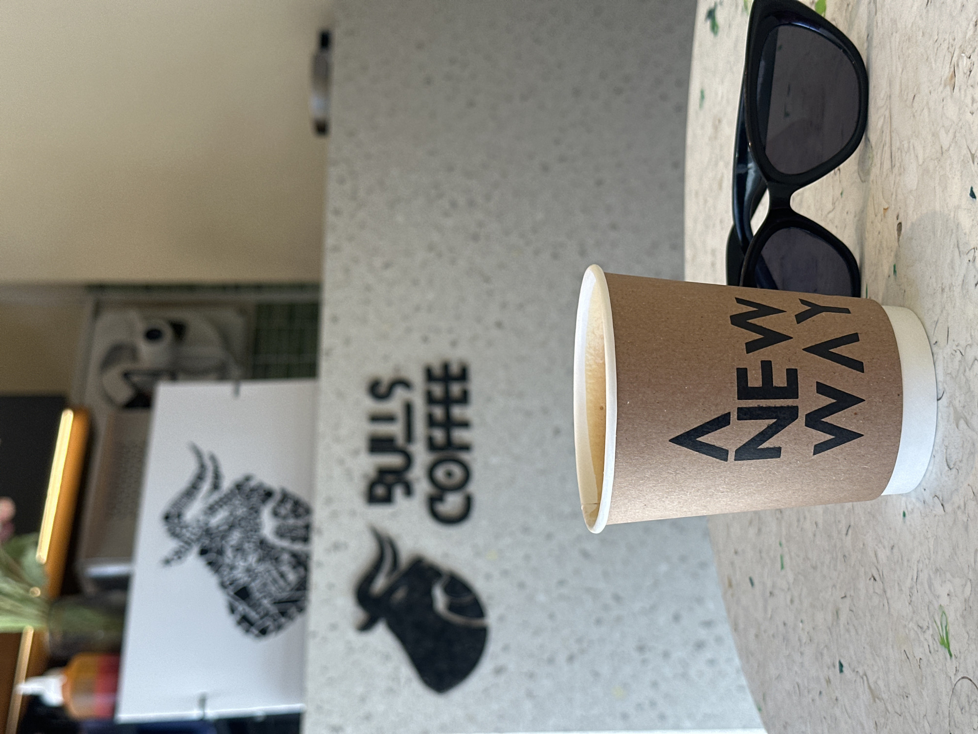 The image depicts a coffee cup, sunglasses, and marketing material in North Kuta, Indonesia.
The coffee cup is cylindrical, with a light brown exterior and a white rim. The words "A New Way" are printed vertically on the side. To the right of the cup are black sunglasses. To the left is marketing material for "Bulls Coffee". A silhouette of a bull appears on the graphic, in black. The background has a mottled, light gray surface, typical of a cafe table or countertop. The scene is lit with natural light.