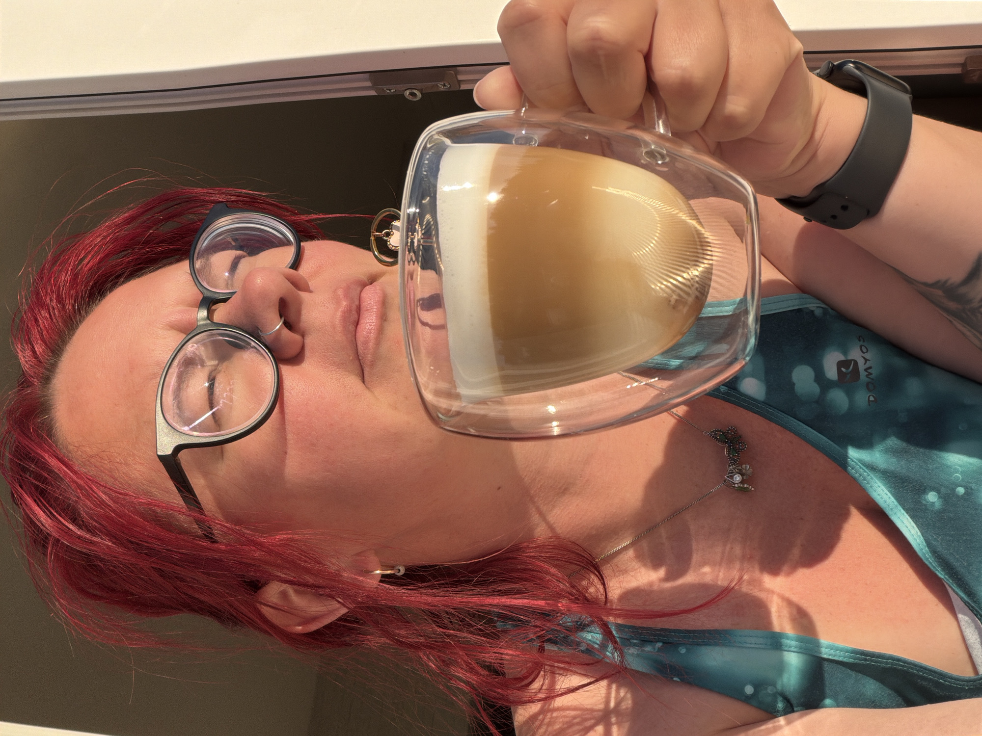 A person with red hair, wearing eyeglasses and a smart watch, is seen holding a clear glass mug filled with a layered coffee drink. The drink appears to be a latte, cappuccino, or similar beverage, with visible layers of milk or foam and coffee. The person is wearing a light green tank top with white circles on it. The photo seems to be taken indoors, possibly near a window, as indicated by the light source and the window frame visible in the background. No information from the image confirms the location to be Chaikovsky, Russia.