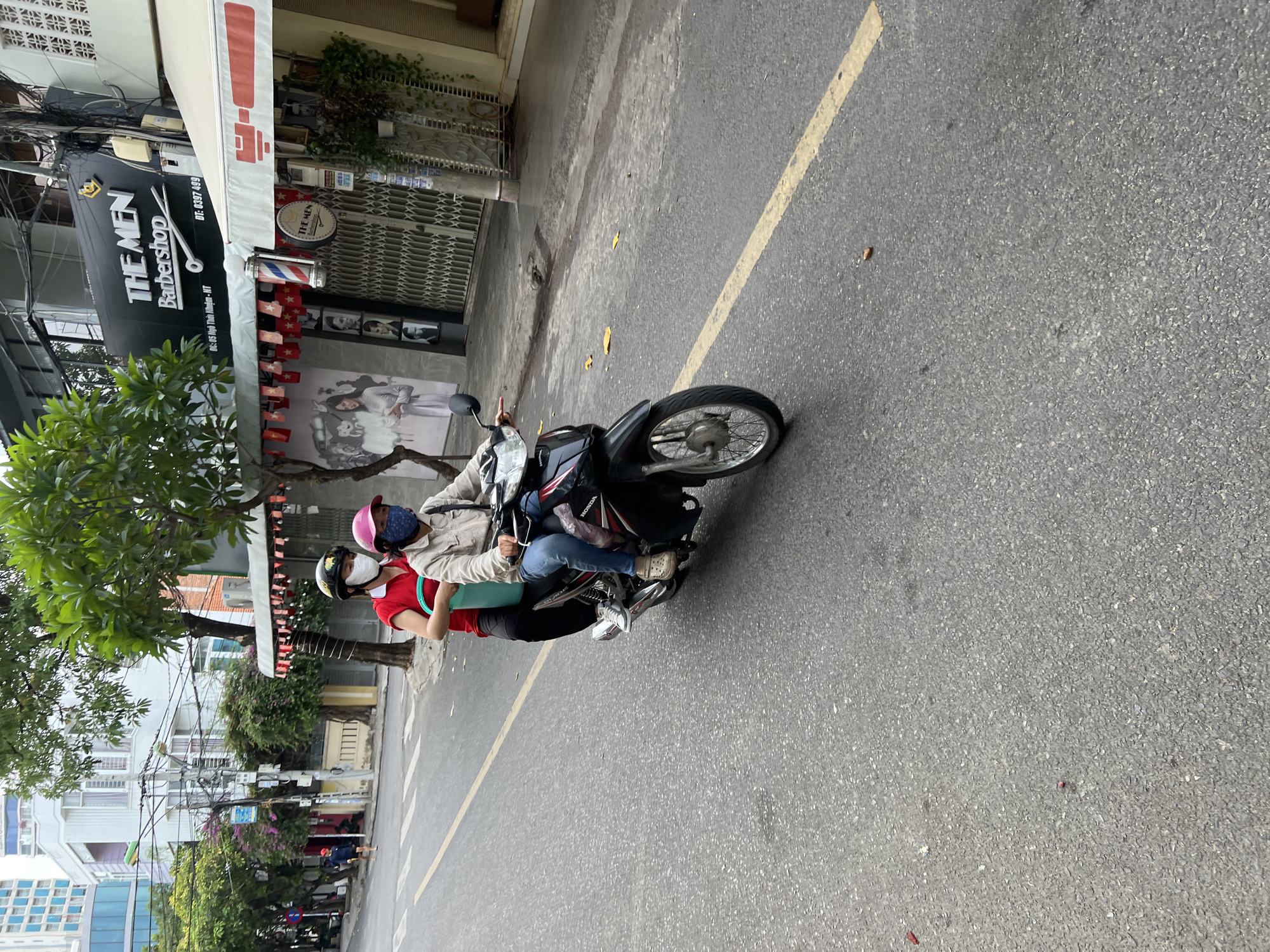 In Nha Trang, Vietnam, two individuals are riding on a black Honda motorcycle on a paved city street. The person in the front, wearing a pink helmet and a light-colored jacket over a dark shirt, is operating the vehicle. The person behind, wearing a black helmet and a red shirt, is seated on the back and holding a light blue bucket. Both individuals are wearing face masks. The motorcycle is traveling on the right side of the road, marked by a yellow line. In the background, a building with signs for "THE MEN Barbershop" is visible, along with other businesses and trees. The environment appears to be a commercial street in an urban area.