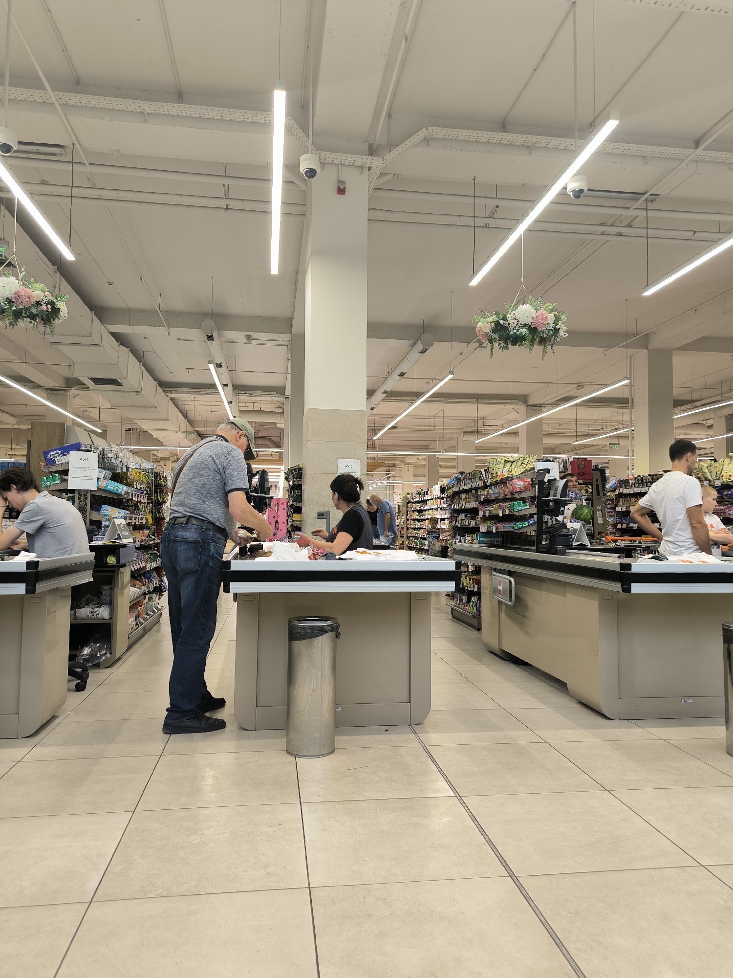 The image depicts a checkout area within a supermarket in Chișinău, Moldova. The scene is brightly lit by overhead fluorescent lights and features multiple checkout lanes with light-colored counters. In the foreground, a man wearing a grey t-shirt and jeans is standing at a checkout counter, interacting with a female cashier who is wearing a black t-shirt. The man appears to be handing items to the cashier. To the left, another man in a grey t-shirt is seated at a checkout, seemingly engaged with his phone. Further down the checkout lanes, other individuals are visible, including a man in a white t-shirt standing at a counter. Shelves stocked with various products line the background, typical of a retail environment. Decorative floral arrangements hang from the ceiling. The floor is tiled in a neutral color. Security cameras are visible on the ceiling.