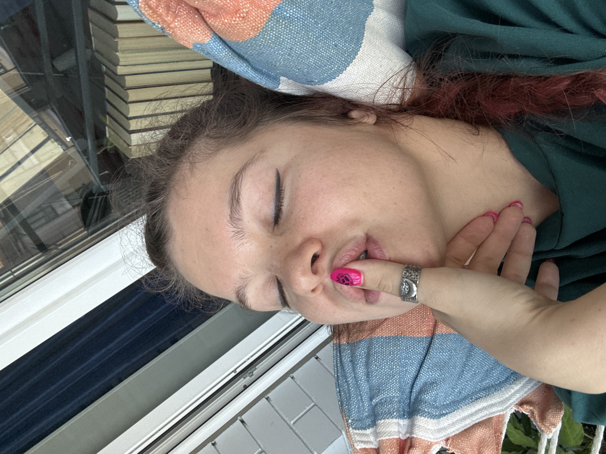 A close-up, portrait-oriented image shows a young person resting or sleeping indoors in Vladimir, Russia. The individual, appearing to be female, is depicted from the chest up, lying on their side with eyes gently closed, conveying a sense of peace and relaxation. They have light skin, dark hair with visible reddish/pinkish highlights, and are wearing a dark green top.
The person's left hand, adorned with bright pink nail polish featuring a black design (possibly a paw print) on the index finger, is held near their mouth, with the index finger lightly touching or slightly inside the lips. A detailed silver ring, possibly depicting a crescent moon and other symbols, is worn on the same finger.
In the background, a tall stack of books is visible, neatly piled, suggesting a setting conducive to reading or study. A colorful striped blanket or towel, featuring patterns of orange, blue, and white, is draped around the person's head and shoulder area. Through a white-framed window, natural daylight illuminates the scene, indicating it is daytime. Part of an outdoor view or blinds/curtains are visible through the window. There is no visible text in the image.