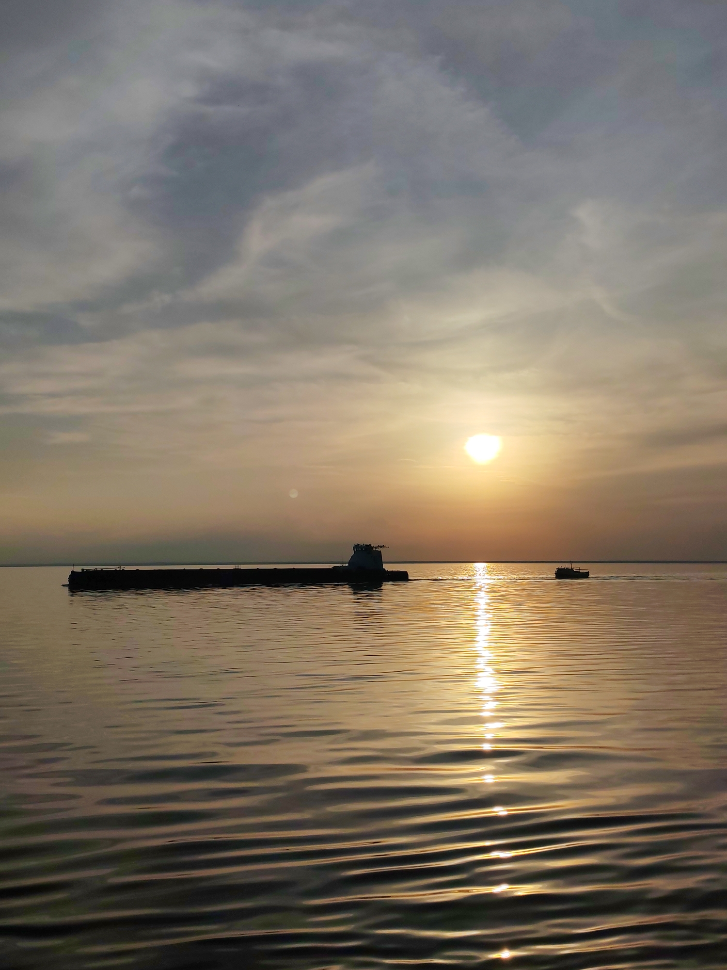 The image depicts a sunset scene in Belozersk, Russia. A large, dark-colored cargo ship is silhouetted against the setting sun. The sun reflects on the calm waters of a lake or river, creating a bright, vertical line of light. A smaller boat is visible further in the distance, heading towards the horizon. The sky is covered with light, wispy clouds, adding to the sunset atmosphere.