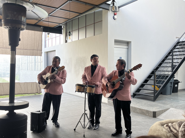 Three men are performing music in a modern, open-air space. They wear matching pink blazers and black pants. One plays a guitar, another a smaller stringed instrument, and the third is on percussion, playing small drums. A tall patio heater stands to the left, and a black metal staircase is visible on the right. The lighting is bright, and the scene appears to be part of an event or performance.