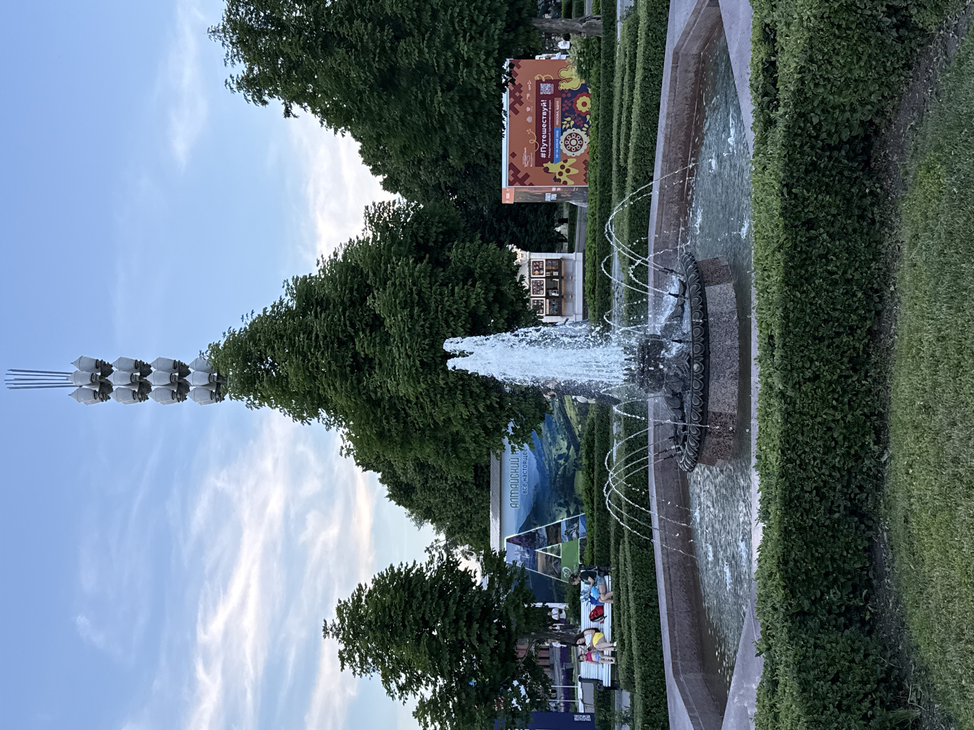 The image captures a public park scene in Moscow, Russia, featuring a central fountain with water spraying upwards. The fountain is surrounded by manicured hedges and a paved pathway. In the background, several large trees provide shade. To the left, a banner with the text "Алтайский Край" and images of mountains and industrial structures is displayed. Further left, people are seated on a bench, enjoying the park. To the right, another banner with the text "#Путешествуй!" and decorative patterns is visible, along with a small structure. At the far end of the park, a tall, slender tower topped with multiple light fixtures stands against a cloudy sky. The overall atmosphere suggests a pleasant, possibly late afternoon or early evening, setting in a well-maintained urban park.