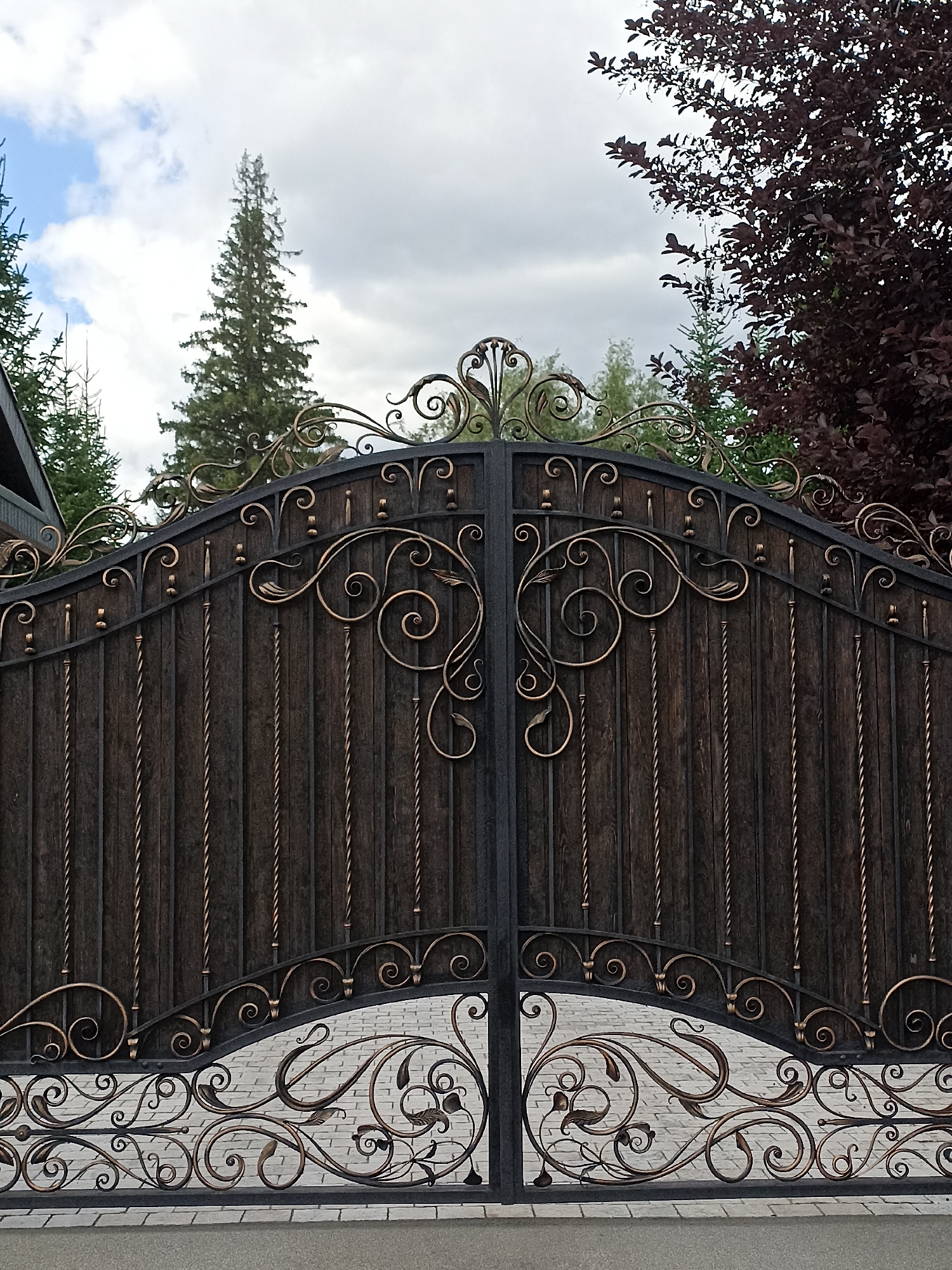 A pair of ornate, dark brown wooden gates with wrought iron accents are shown in the foreground. The gates feature intricate scrolling metalwork in a gold tone, with decorative finials and twisted metal uprights. Behind the gates, a cobblestone driveway is visible, and beyond that, a lush green landscape with tall pine trees and a dense, dark red tree. The sky above is partly cloudy, with patches of blue sky and white clouds. The overall scene suggests a well-maintained property entrance. The image is captured from a frontal, slightly low-angle perspective.