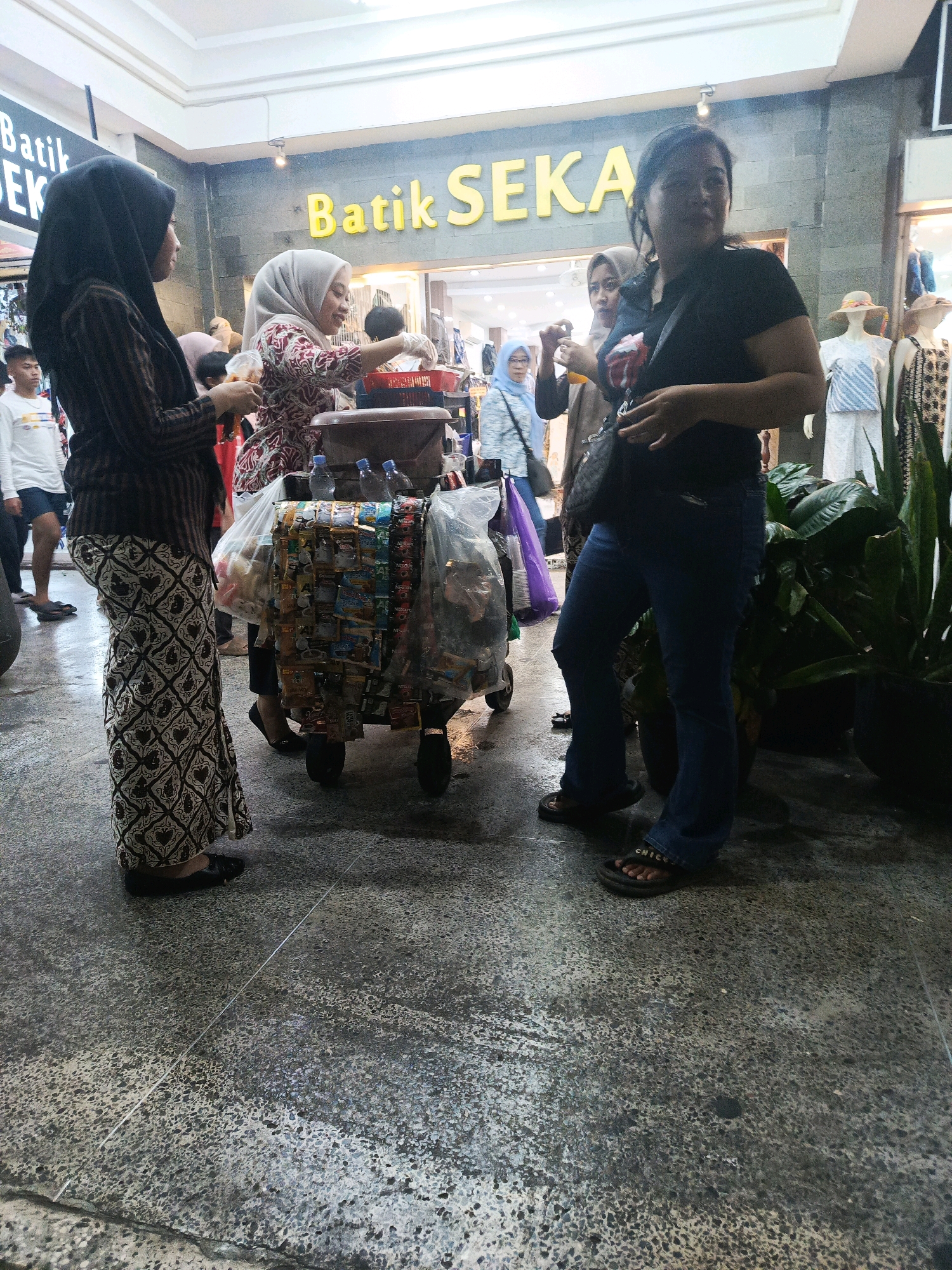 An outdoor scene in what appears to be a marketplace or shopping area in Yogyakarta, Indonesia, features a group of people gathered around a mobile cart selling various items, likely snacks or small goods. A woman in a black hijab and a patterned skirt stands to the left, facing the cart. Beside her, a woman in a light-colored hijab and a patterned top is interacting with the cart, possibly making a purchase. Further to the right, a woman in a black t-shirt and blue jeans is looking towards the camera and appears to be holding a bag. In the background, mannequins display clothing in a shop window, and signage for "Batik SEKA" is visible. The ground is paved with textured concrete. The lighting suggests it is daytime.