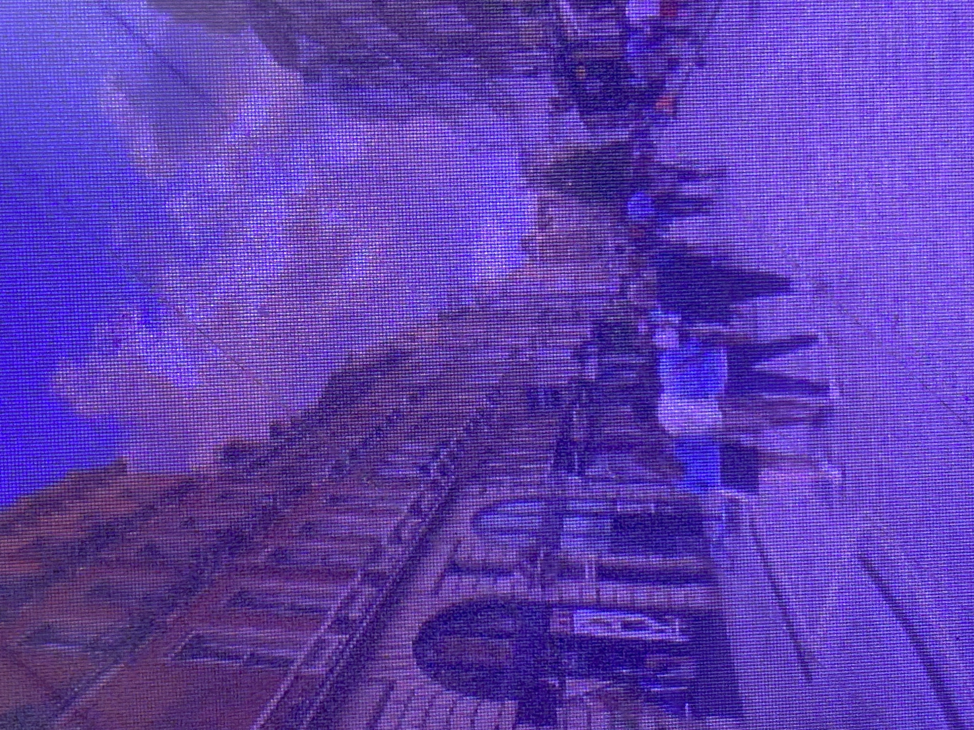 The image depicts a street scene in Ikorodu, Nigeria, illuminated by a pervasive purple hue. The architecture on the left suggests a European influence, with a multi-story building featuring arched windows and visible brickwork. The street is paved, and several figures are walking along it. In the foreground, four individuals are visible, two wearing white tops and dark bottoms, and two in darker clothing. Further down the street, more people are present, appearing as silhouettes against the illuminated sky and buildings. The sky is a deep purple with hints of lighter clouds. The overall impression is of a city street at dusk or under artificial purple lighting.