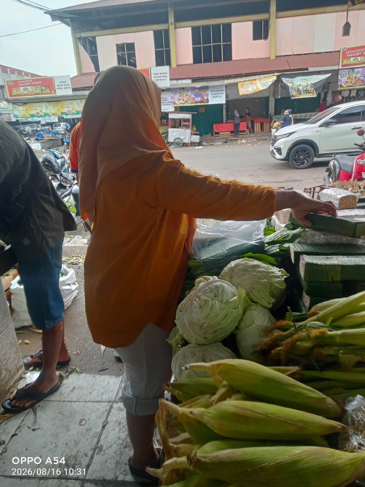 In Jambi, Indonesia, this image captures a street market scene. A person wearing an orange hijab and a yellow shirt is visible from the side, reaching towards a display of fresh produce. The foreground features an abundance of corn on the cob and cabbage heads, suggesting a vibrant food market. Behind the produce, several white tempeh blocks wrapped in banana leaves are stacked neatly. In the background, a white SUV is parked, and a red scooter is visible to the right. The market appears to be under a covered structure with shops lining the street. The overall atmosphere suggests a typical day at a local Indonesian market.