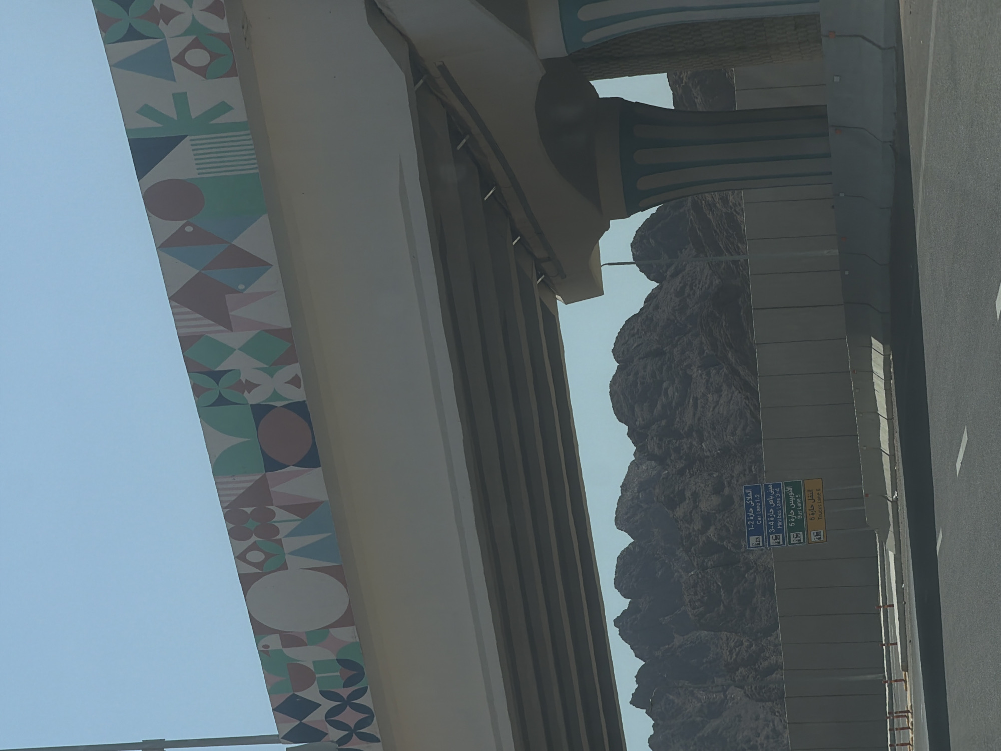 The image shows a multi-level highway overpass with decorative geometric patterns on the underside of the upper level. The overpass is supported by large, fluted columns that are painted with alternating light blue and white vertical stripes. In the background, there are rocky, mountainous formations under a clear blue sky. To the right of the image, traffic signs are visible on a concrete barrier, indicating lane designations for cars, mini-buses, buses, and trucks, with text in both Arabic and English. The presence of these road signs and the modern infrastructure suggest an urban or semi-urban setting. Given the style of the architecture and the mountainous backdrop, this scene could be located in an area like Sharm El Sheikh, Egypt, known for its coastal resorts and mountainous terrain. The image is taken from the perspective of someone traveling on a road, looking upwards at the overpass and the surrounding landscape.