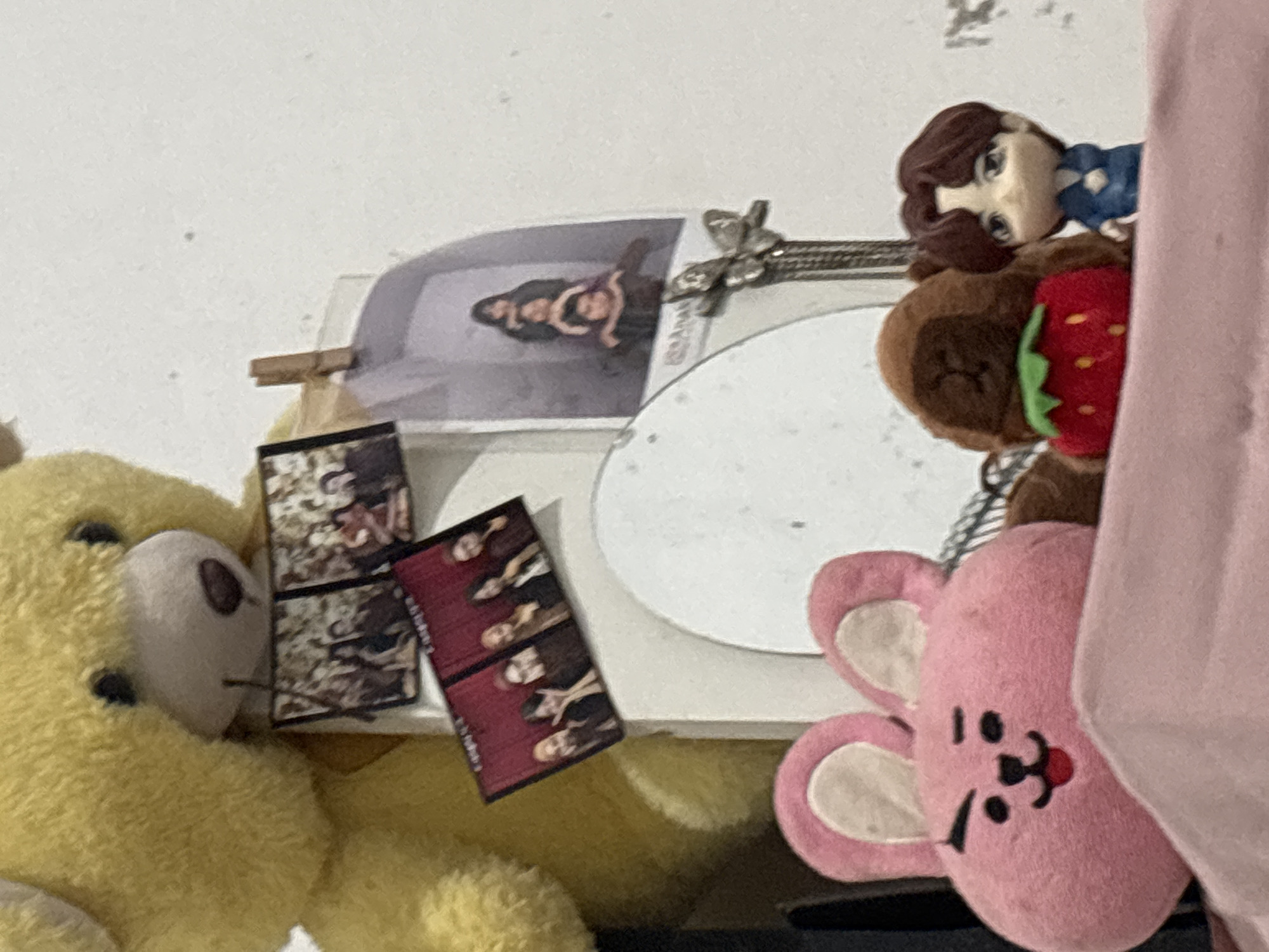 A close-up, indoor, eye-level shot focuses on a collection of plush toys and small decorative items displayed against a plain white wall. In the foreground, a pink bunny-shaped plush toy with black embroidered features, including eyes, eyebrows, a nose, and an open mouth with a red tongue, is partially visible. Next to it is a small, brown, round plush toy with a red strawberry design on its front and a green stem. To the right of the strawberry toy is a small, stylized doll with dark hair, large blue eyes, and wearing a blue outfit.
Behind these plush toys, a large, fuzzy yellow teddy bear is visible, primarily its head and upper torso. Attached to the teddy bear or leaning against it are several small photographs or postcards. Two are clustered together, showing groups of people. Another photograph, clipped to a wooden clothespin and leaning against a white framed object, depicts four individuals stacked vertically. To the left of this, another set of two smaller photographs shows groups of people. A large, circular, white object, possibly a mirror or a decorative plate, is positioned behind the plush toys. Further to the right, a metallic butterfly-shaped ornament hangs from a chain. The overall impression is of a curated display of personal items within a domestic setting. The image does not contain information that would allow for the determination of its specific location in North Jakarta, Indonesia.