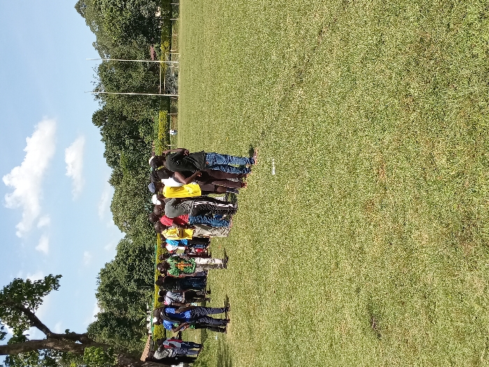 The image captures an outdoor scene on a sports field in Kakamega, Kenya. Approximately 30 to 40 individuals, predominantly African males, are gathered in a cluster on the left side of the frame, standing on a green grass surface at the edge of the field. They are dressed in various casual clothing, including t-shirts, shirts, trousers, and jeans, with some wearing headwear. The group faces generally towards the right, appearing to observe or await an event on the field. The foreground consists of a mown green grass field. In the mid-ground, a set of tall white H-shaped rugby goalposts and a shorter set of football goalposts are visible. A dense treeline forms the background, with a large, mature tree featuring a prominent trunk situated on the far left. A solitary, indistinct white figure is visible in the distant center of the field, near the goalposts. The sky above is blue with scattered white cumulus clouds, and the scene is brightly illuminated by natural sunlight, indicating daytime.
