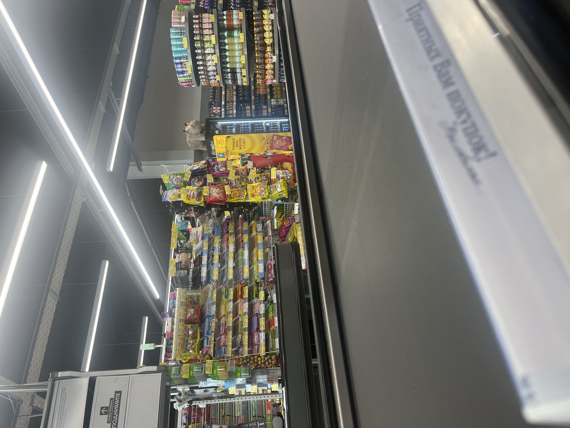 The image shows a checkout counter in a supermarket. On the right side of the image, there are shelves stocked with drinks and snacks. In the center, there is a refrigerated display case with more drinks. On the left side, there are more shelves filled with snacks. The checkout counter has a conveyor belt. The lighting is fluorescent. The scene appears to be indoors. The image is taken from the perspective of a customer standing at the checkout counter. The text on the bottom of the image is in Russian.