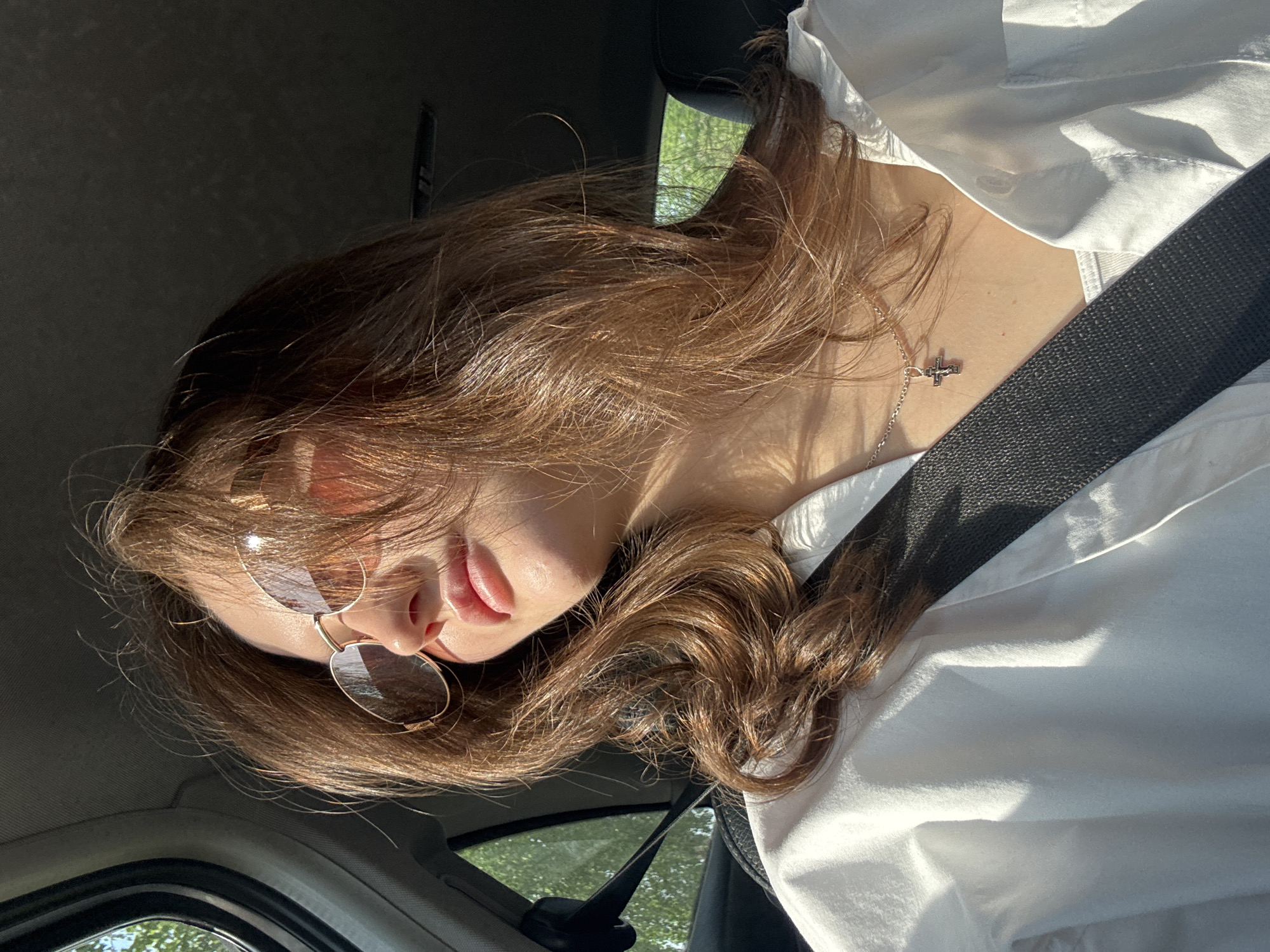A person with brown hair and sunglasses is in a car, wearing a white shirt and a seatbelt. They are facing the camera at an angle. The location is possibly Perm, Russia, but this cannot be confirmed from the image. The person is wearing a necklace with a cross pendant. Sunlight is present in the image. The car's interior is visible.