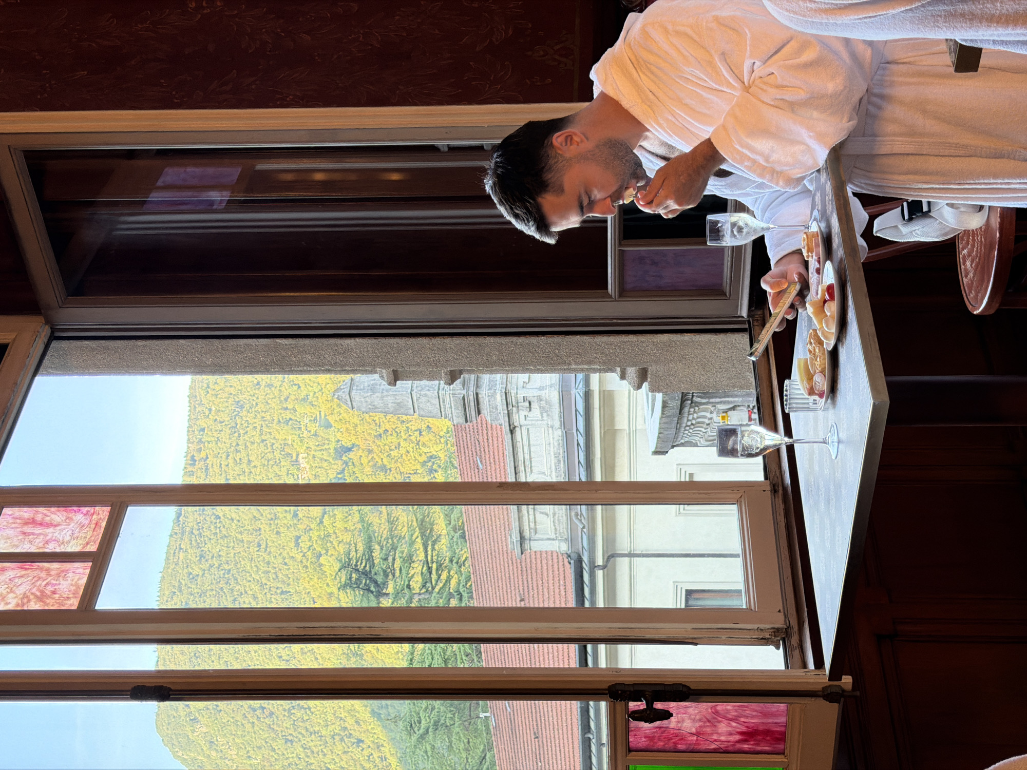A man in a white bathrobe is seated at a table in what appears to be a hotel or spa lobby in San Pellegrino Terme, Italy. He is looking down at his phone and eating. On the table in front of him are two glasses of champagne, a plate with various small pastries, cheese, and some fruit. The table is set against a backdrop of a large window that overlooks a lush, green mountainside with some buildings visible. The window has stained glass inserts in the upper section and a dark wood frame. The interior of the room has dark wood paneling.